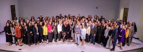 A large, diverse group of people posing for a formal portrait in a brightly lit, spacious room with white walls.
