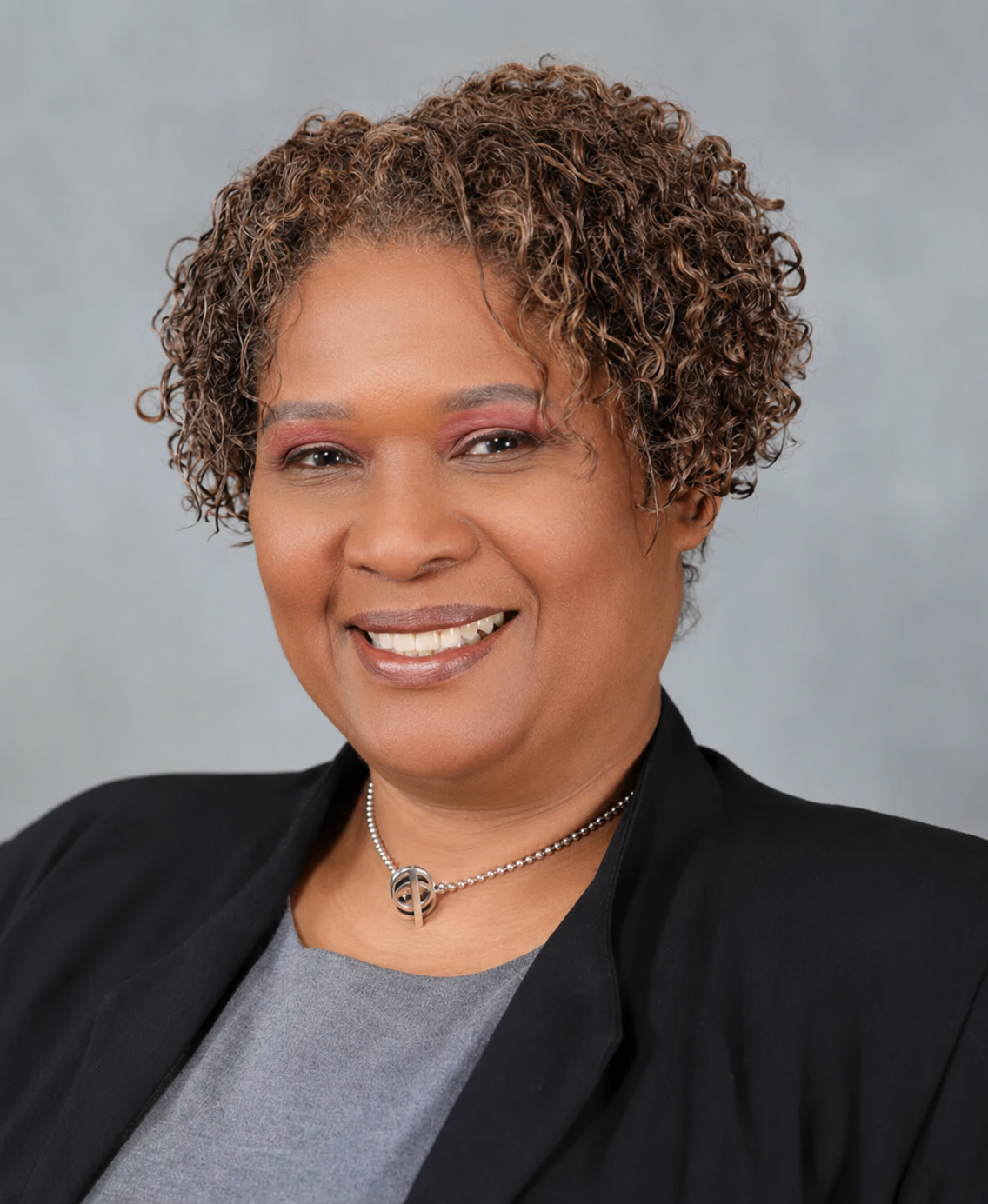 A smiling person with curly hair, wearing a gray shirt, a dark blazer, and a beaded necklace against a gray background.
