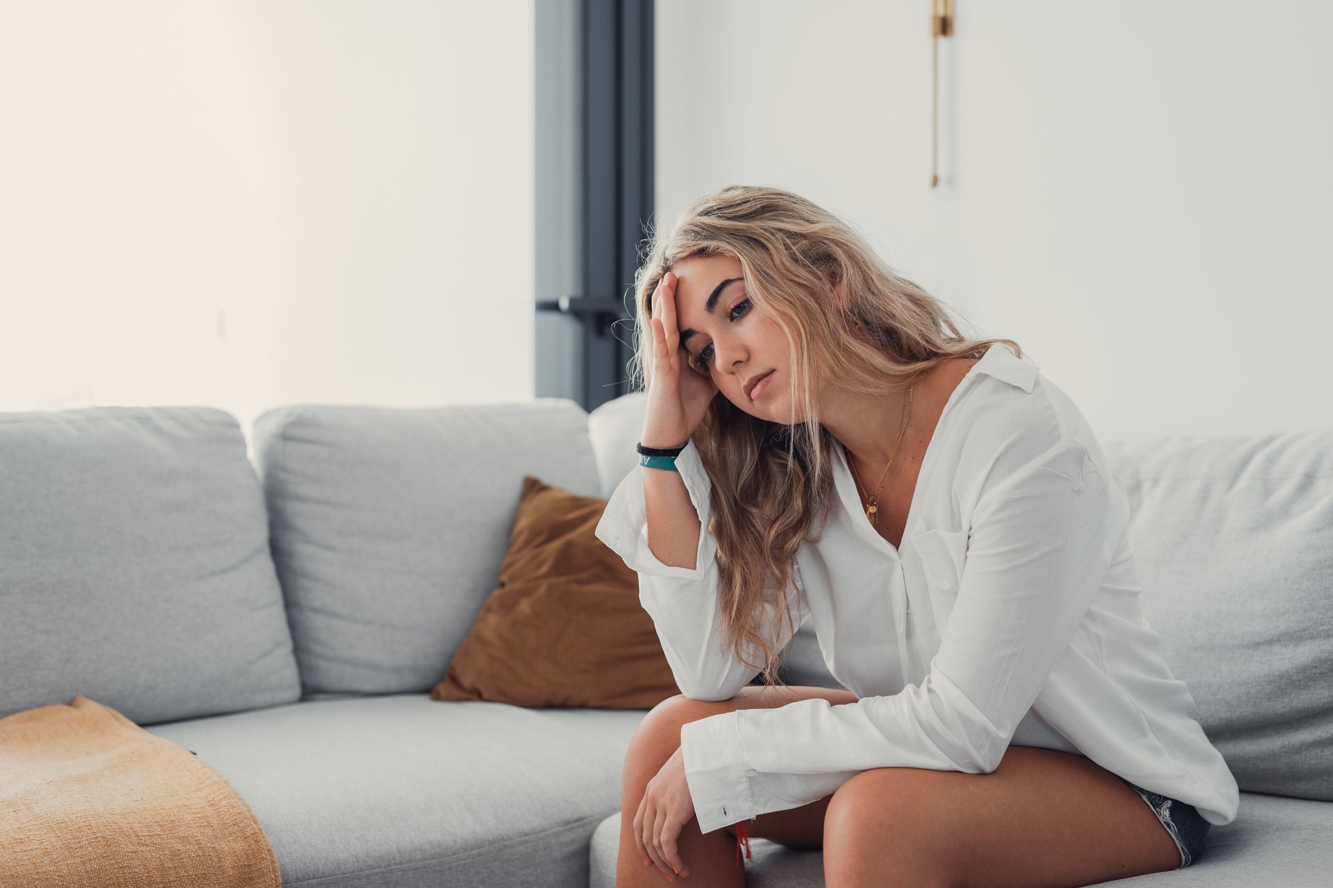 A person with blonde hair sitting on a gray couch, resting their hand on their forehead with a thoughtful expression.