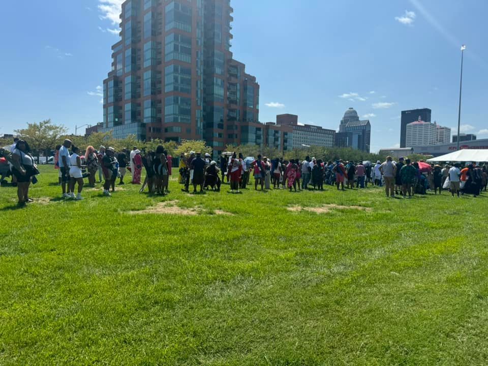 Large crowd gathered outdoors on a grassy area, tall building in the background. Bright sunny day.