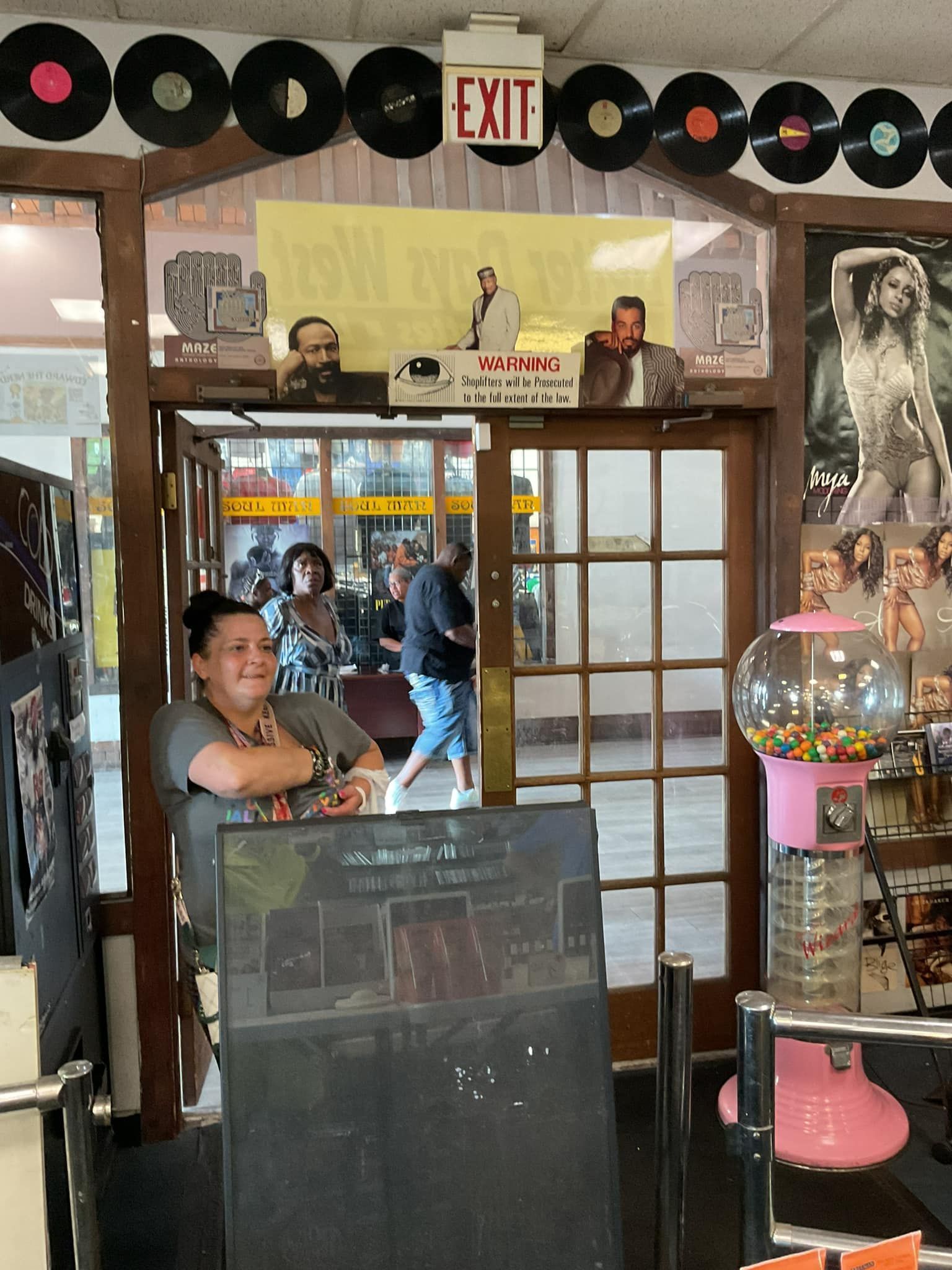 Woman in front of a doorway, looking at camera. Door leads to a hallway. Shop decor with vinyl records and posters.