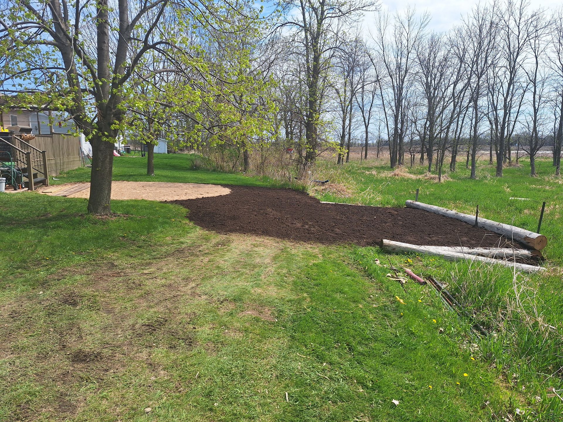 Grassy yard with tree and mulched area, brown and green, with a wooden structure on right.