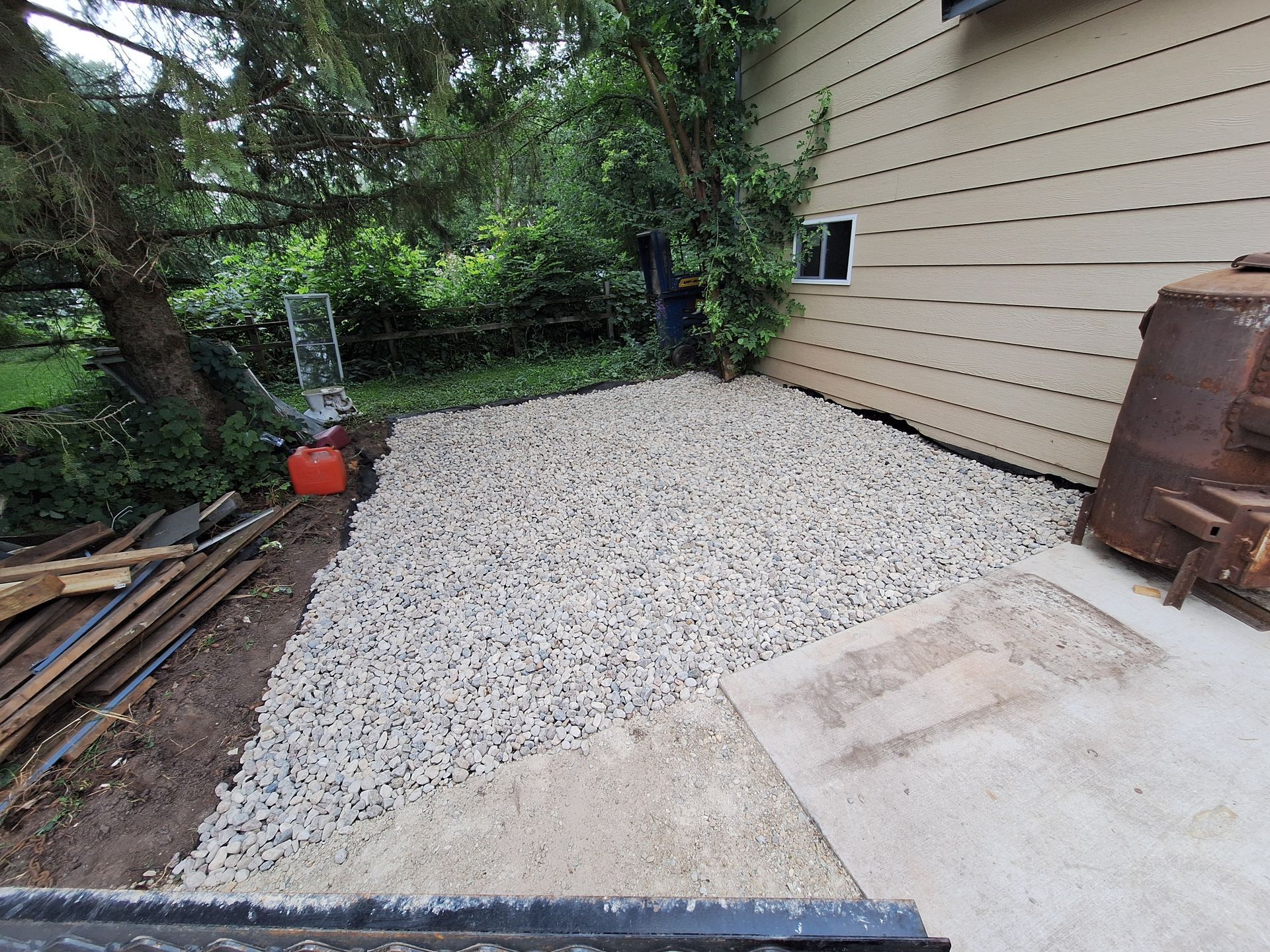 Gravel patio next to house. Concrete steps and siding. Dark green trees.