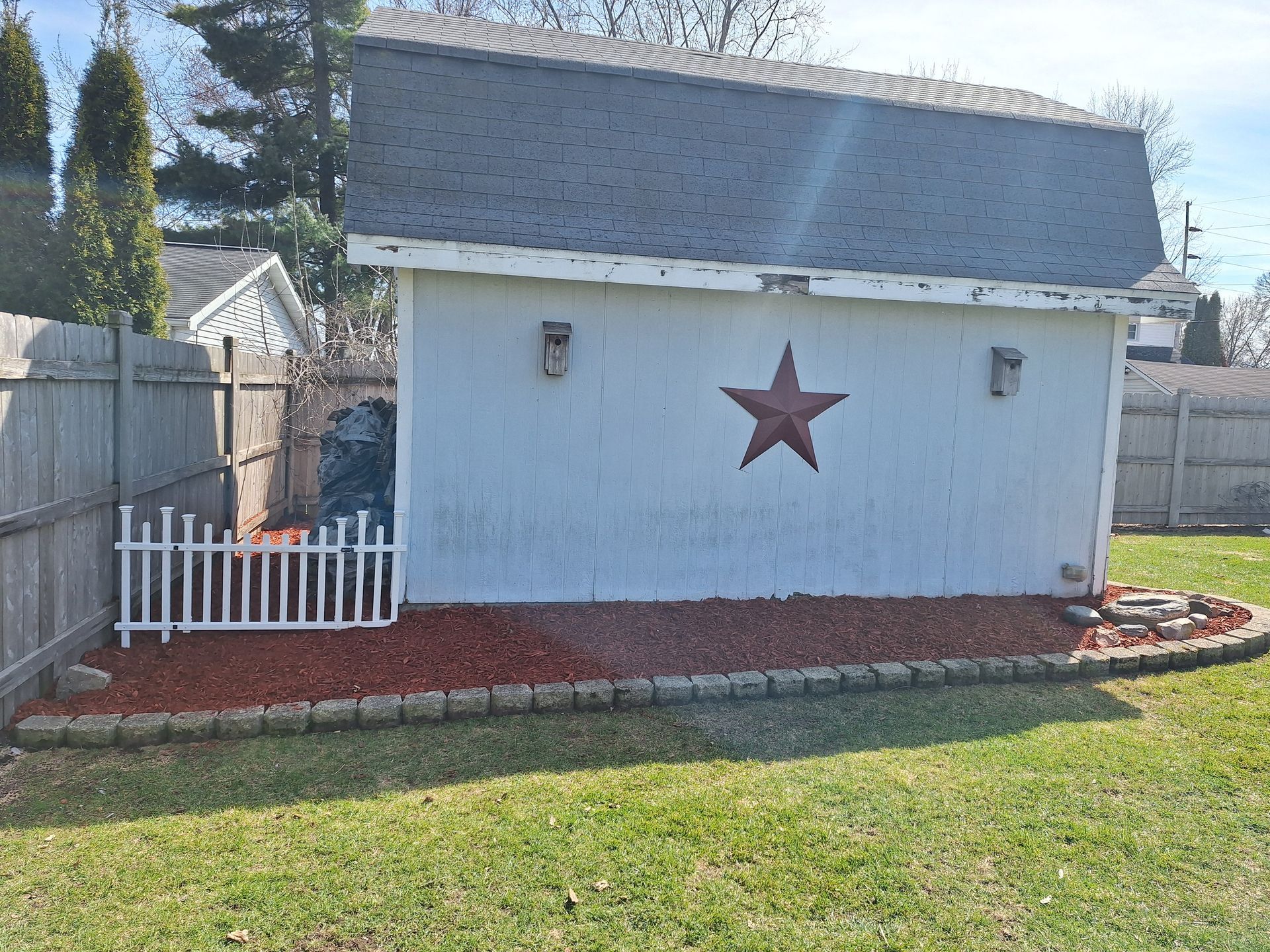 White shed with red star, picket fence, and red mulch border in a grassy yard.