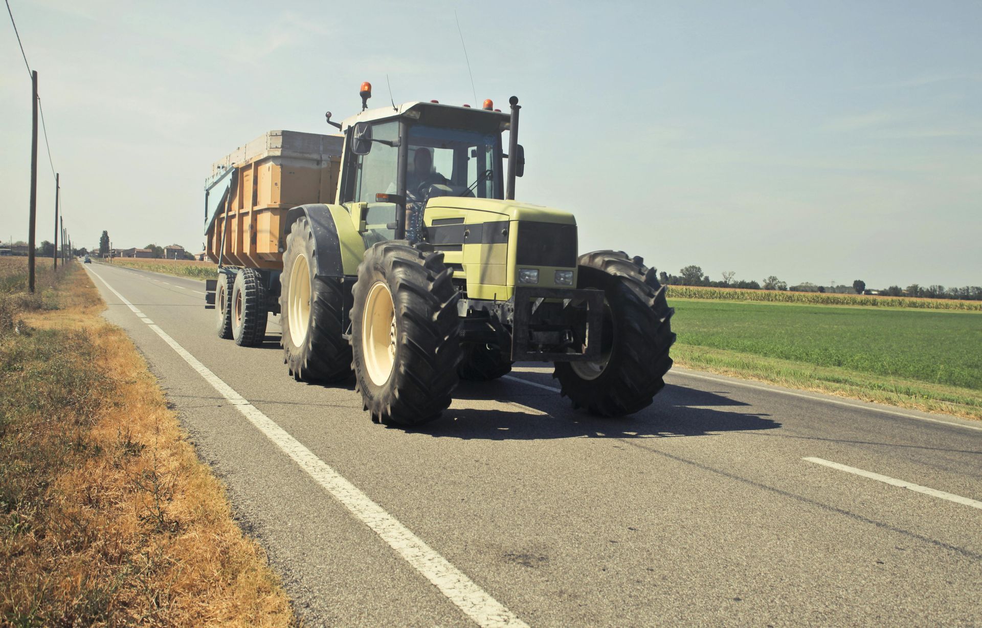 Tractor pulling a trailer on a paved road next to a field on a sunny day.