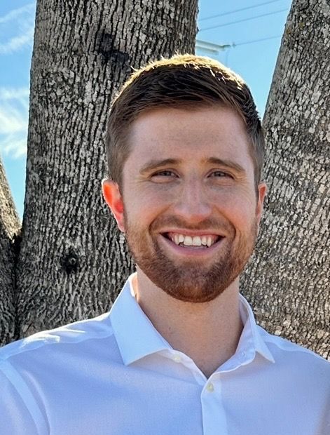 A man with a beard and a white shirt is smiling in front of trees.