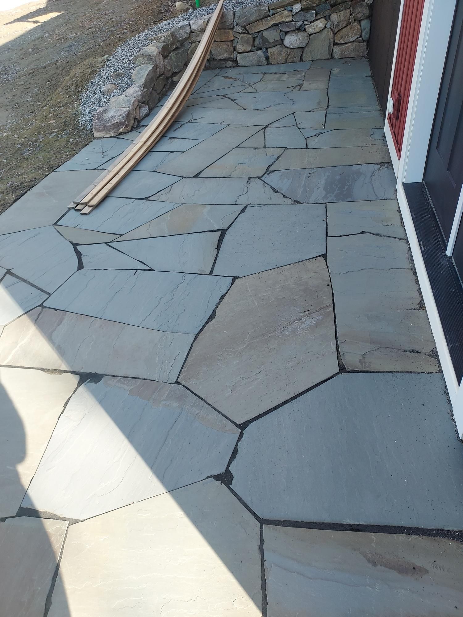 Irregular stone patio in shades of gray with dark grout, next to a red building.