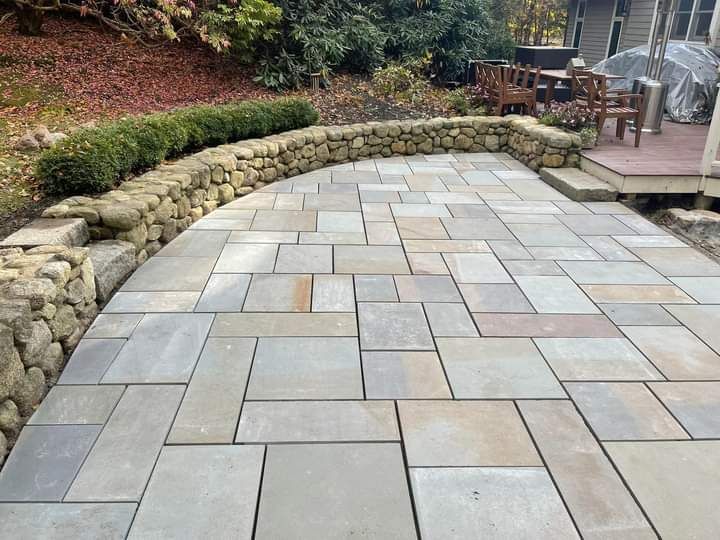 Stone patio with a curved stone wall and bordering greenery. Wooden deck visible in the background.