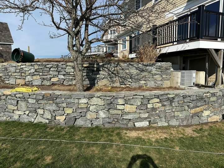 Stone retaining walls with a house and tree. Outdoor setting, sunny day.