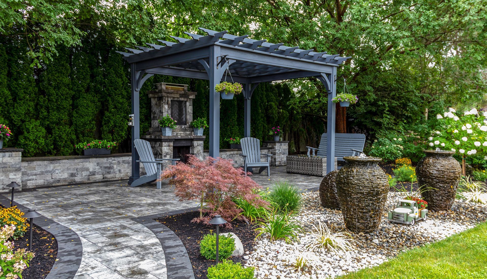 Gray pergola with fireplace and chairs in a garden with stone path, green bushes, and decorative urns.