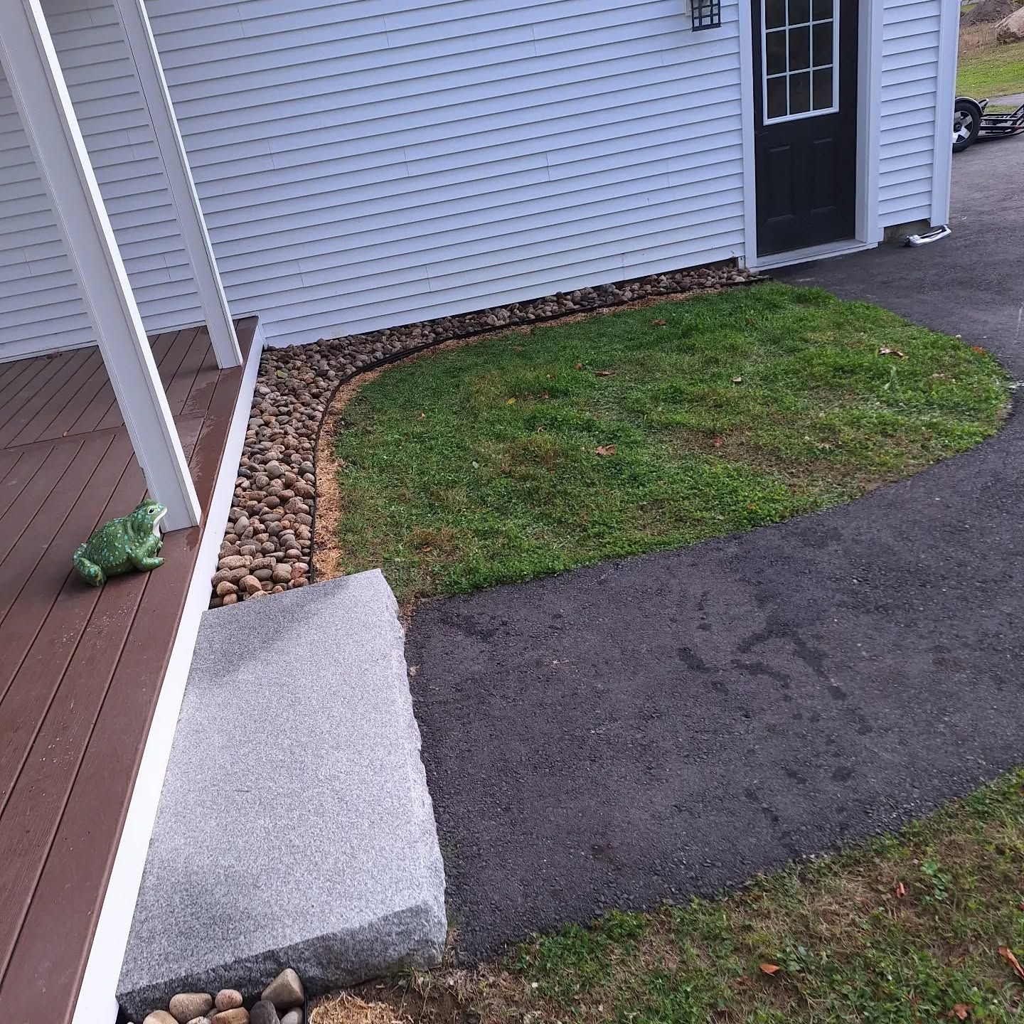 A green frog statue sits on a porch near an asphalt walkway, grass, and a building with a black door.