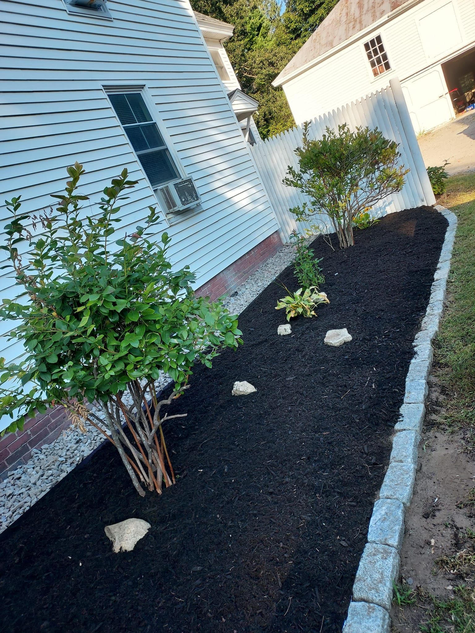 A flower bed with dark mulch and plants, bordered by a stone wall, beside a white house.