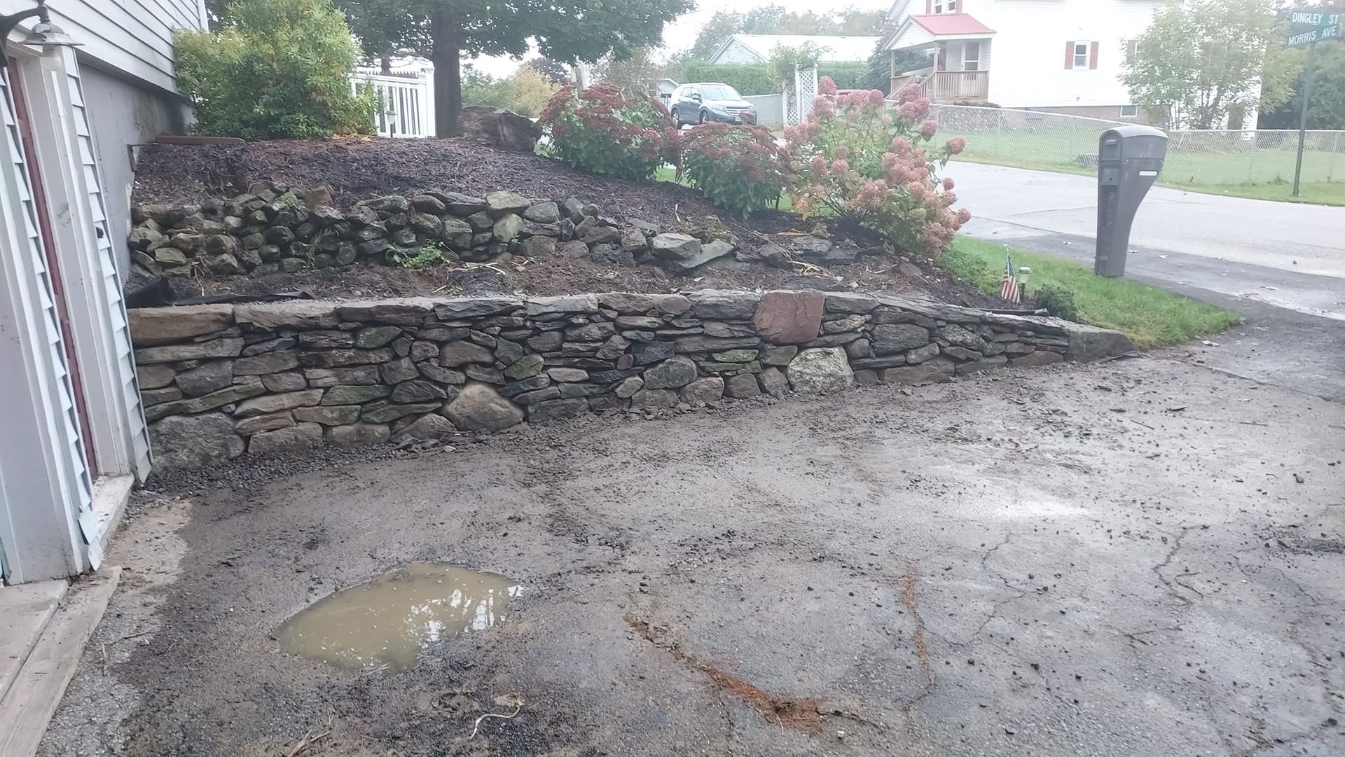 Muddy driveway beside a stone retaining wall with bushes; a house in the background.