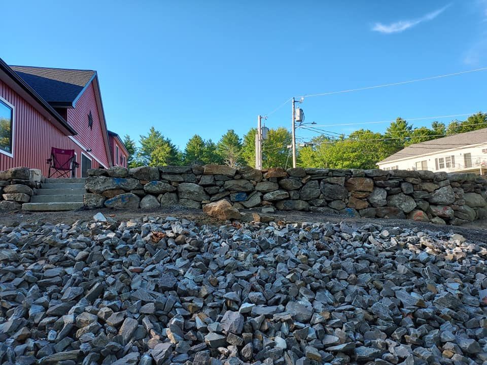 Stone retaining wall in front of a red building and gravel, with trees and blue sky in the background.