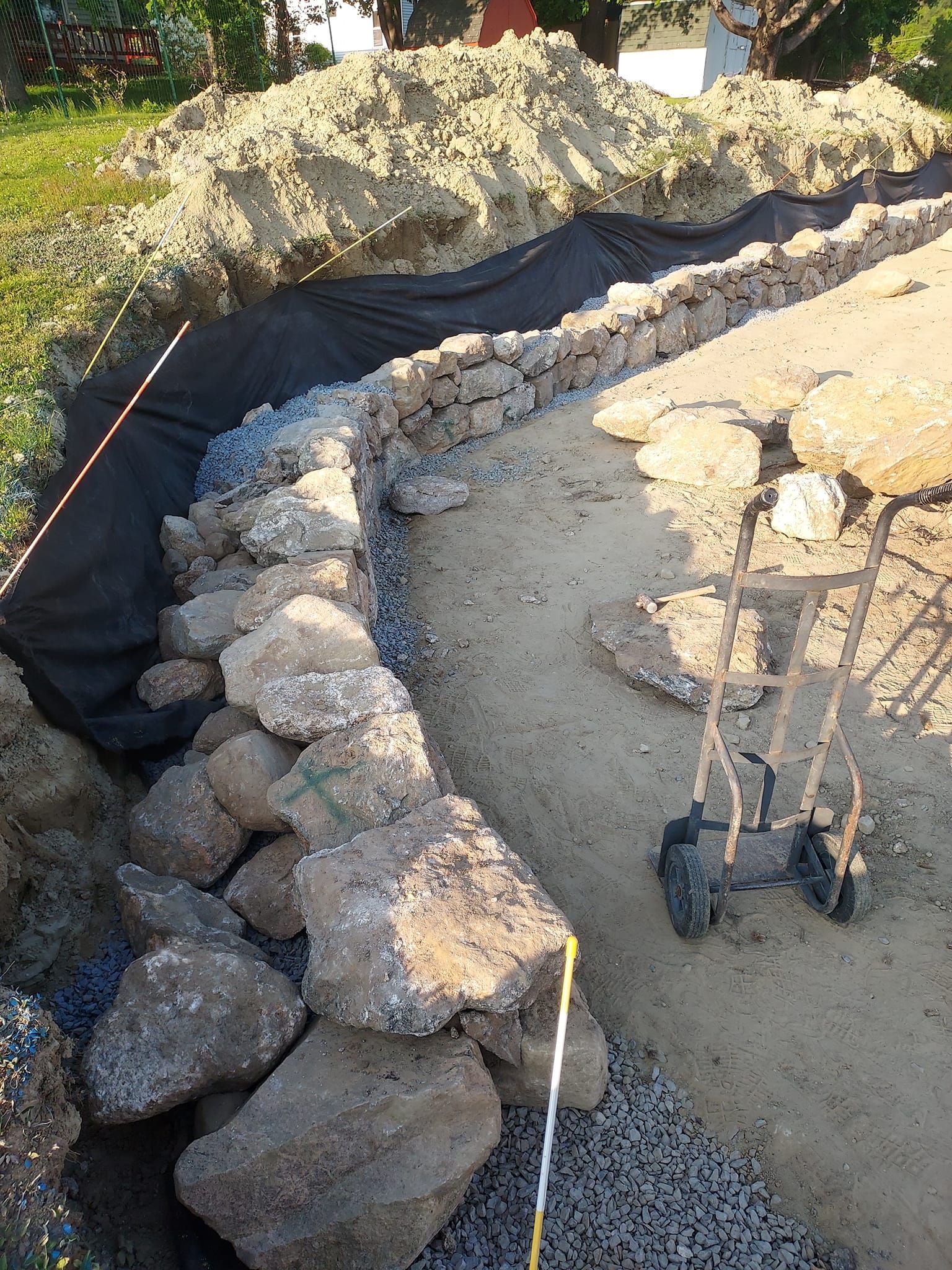 Construction site: rock retaining wall being built along a dirt path. Black fabric liner visible.