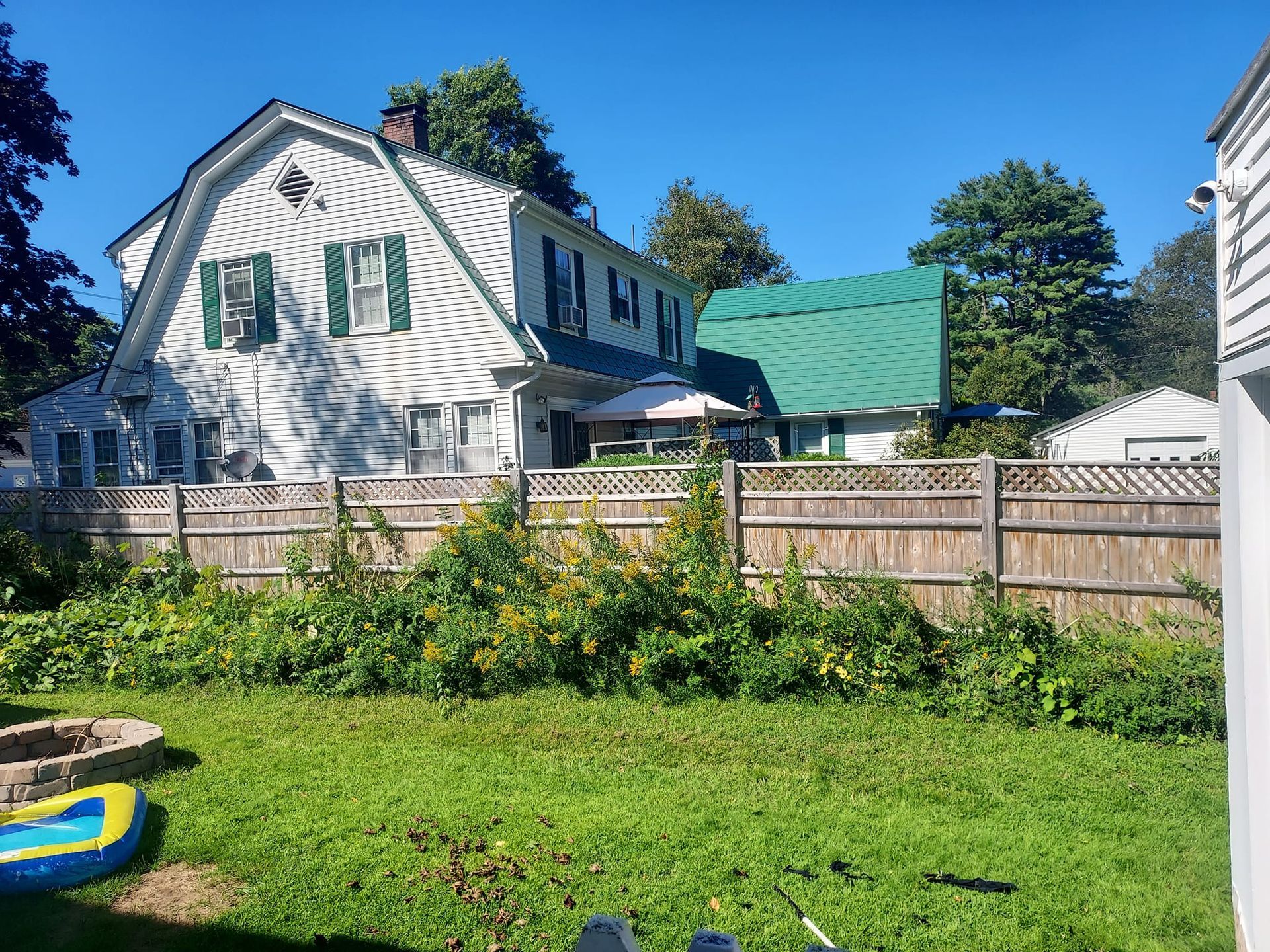 Backyard scene with a two-story white house, green roofed structure, wooden fence, and grassy lawn.