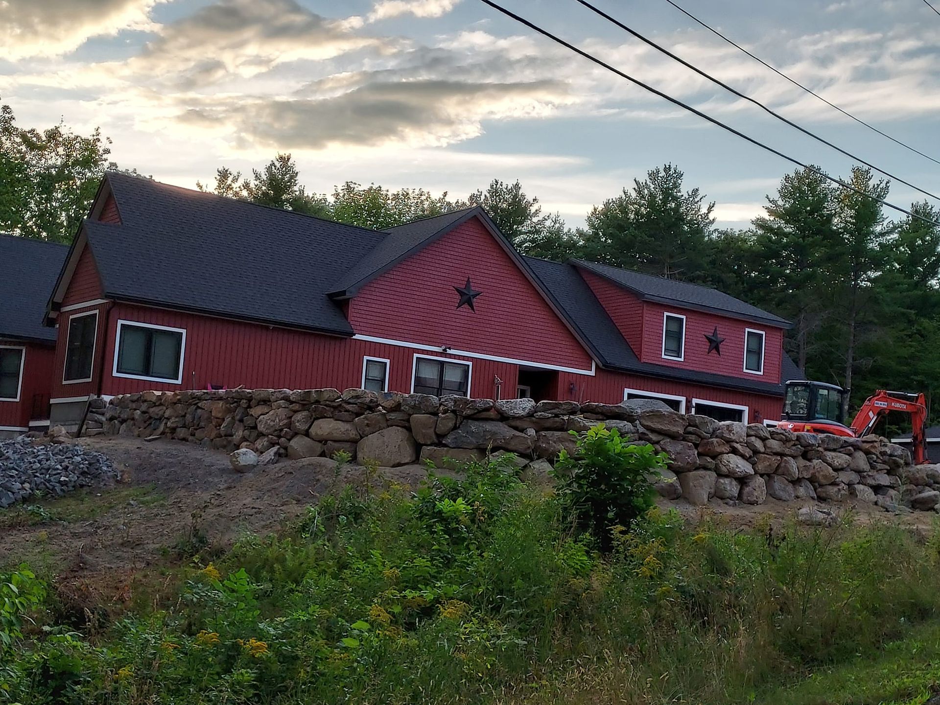 Red barn with black roof, built into a stone wall, with trees and cloudy sky in the background.