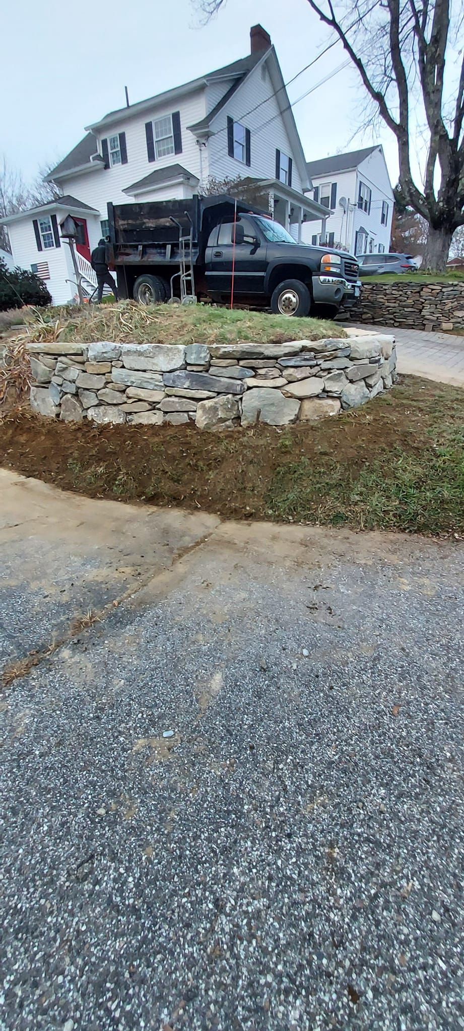 A black dump truck parked near a stone wall with a house in the background. Gravel road.