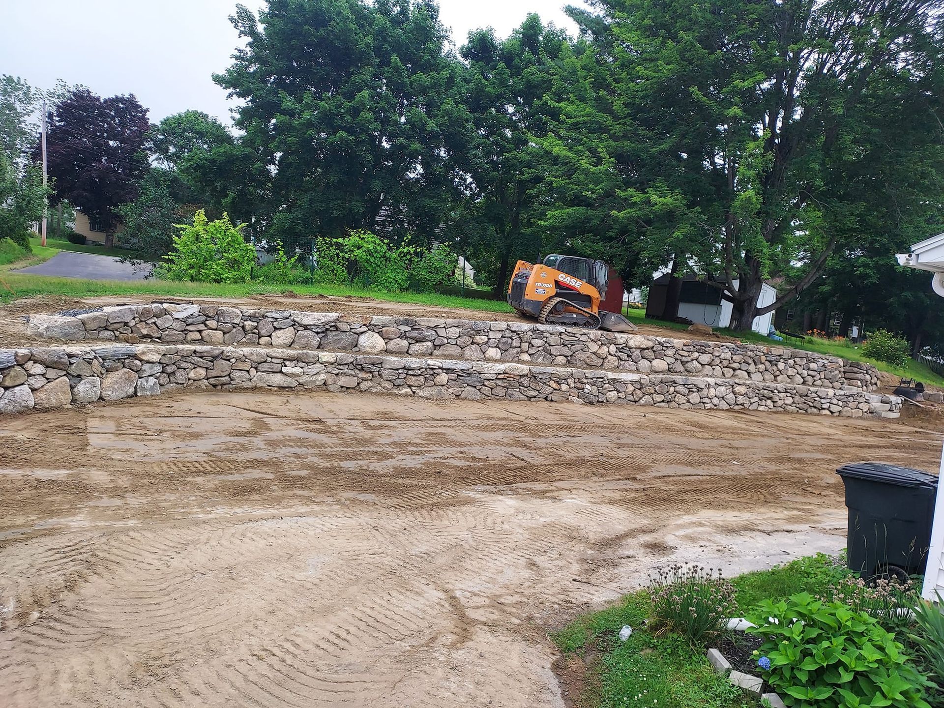 Stone retaining walls on a sloped yard, a small excavator, and bare earth.