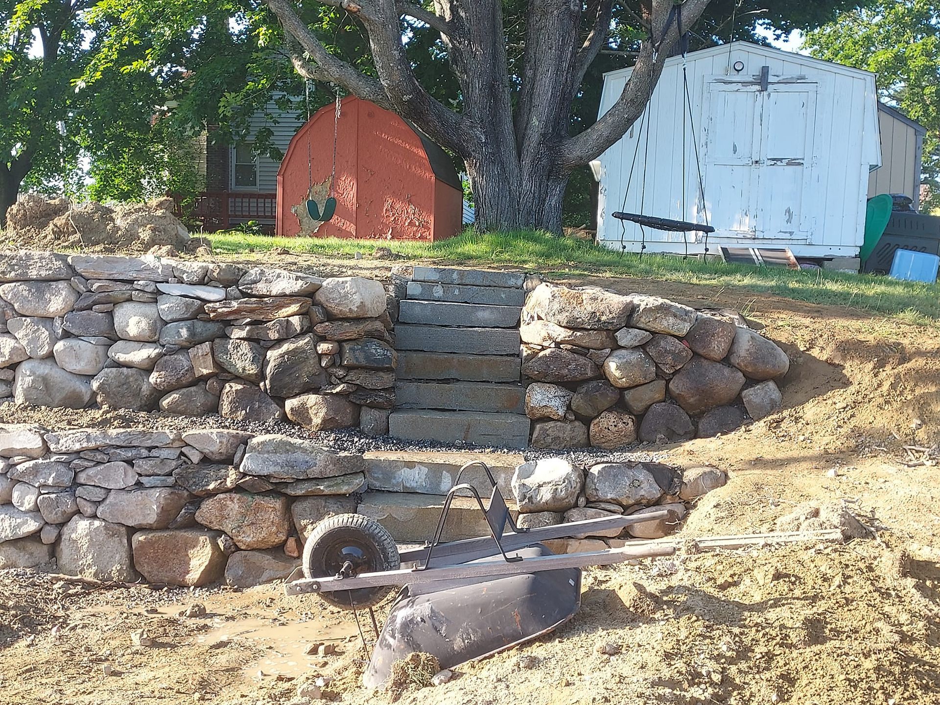 Stone steps and retaining walls in front of red and white sheds, with a garden rake.