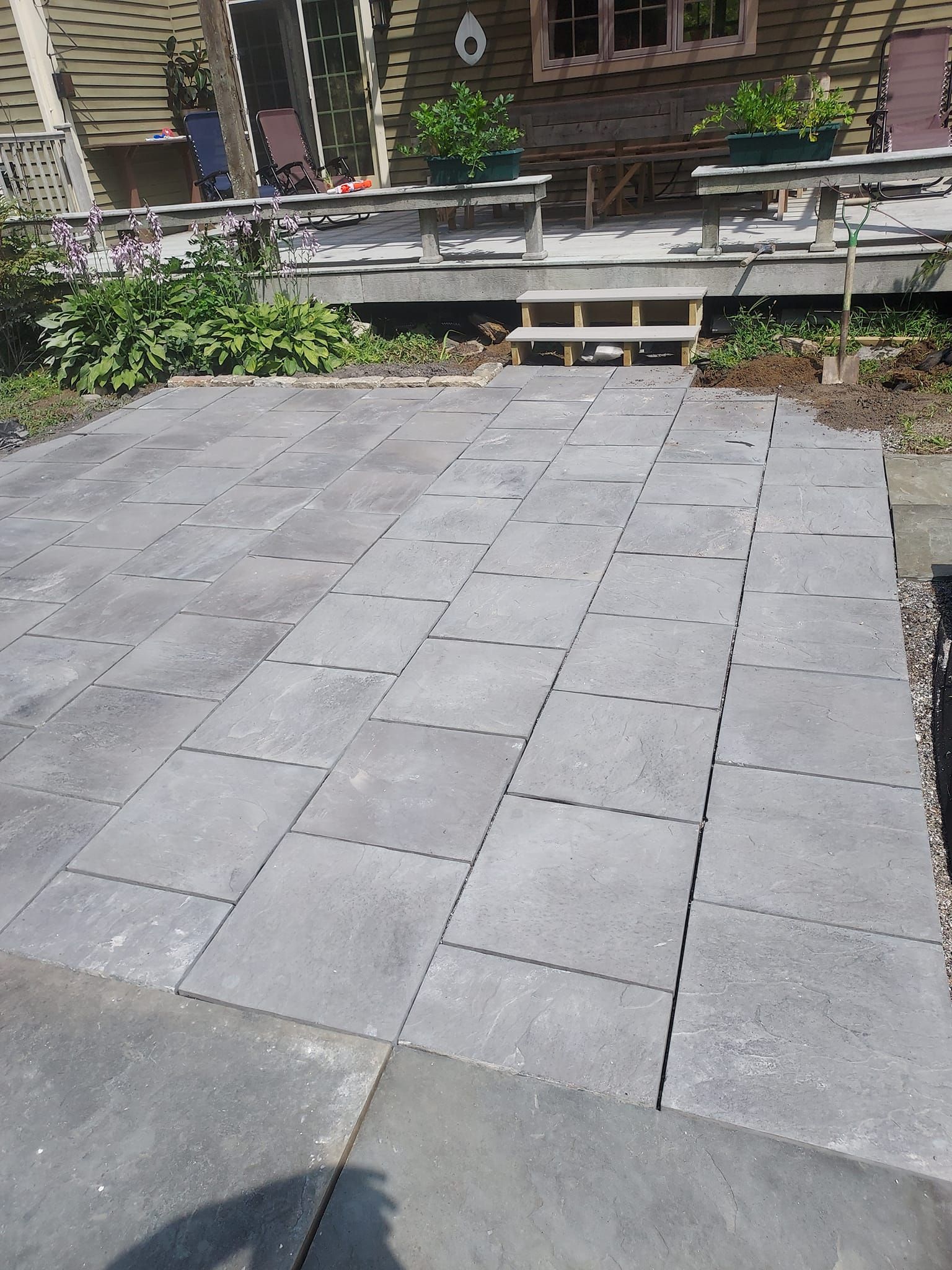 Gray paver patio in front of a house with a wooden deck and steps.