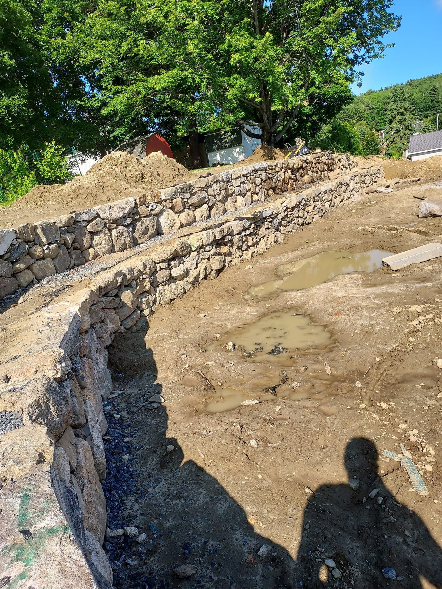 Stone retaining wall under construction, muddy ground, sunny outdoor setting.