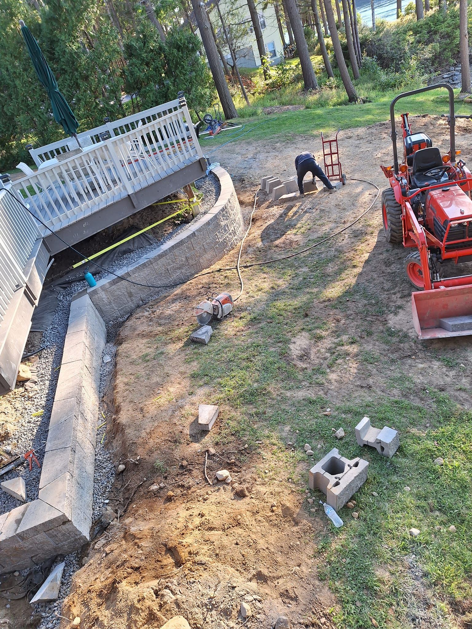 Construction site: person working near a partially built retaining wall under a wooden deck, with a tractor nearby.