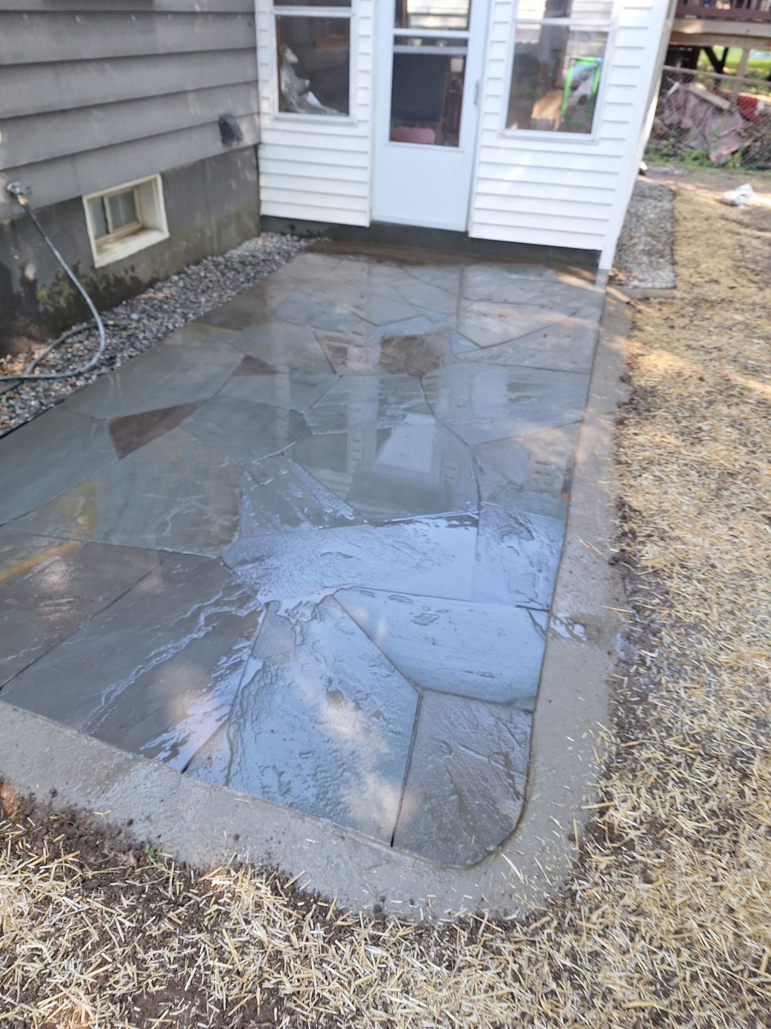 Outdoor patio made of gray stone, next to a white building with a door. Concrete border surrounds the patio.