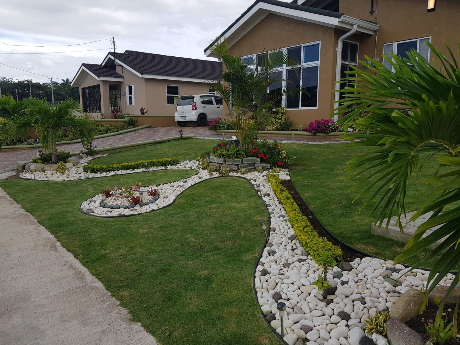 Lush green lawns and flower beds with white stone borders. Two beige houses with white cars parked. Cloudy day.