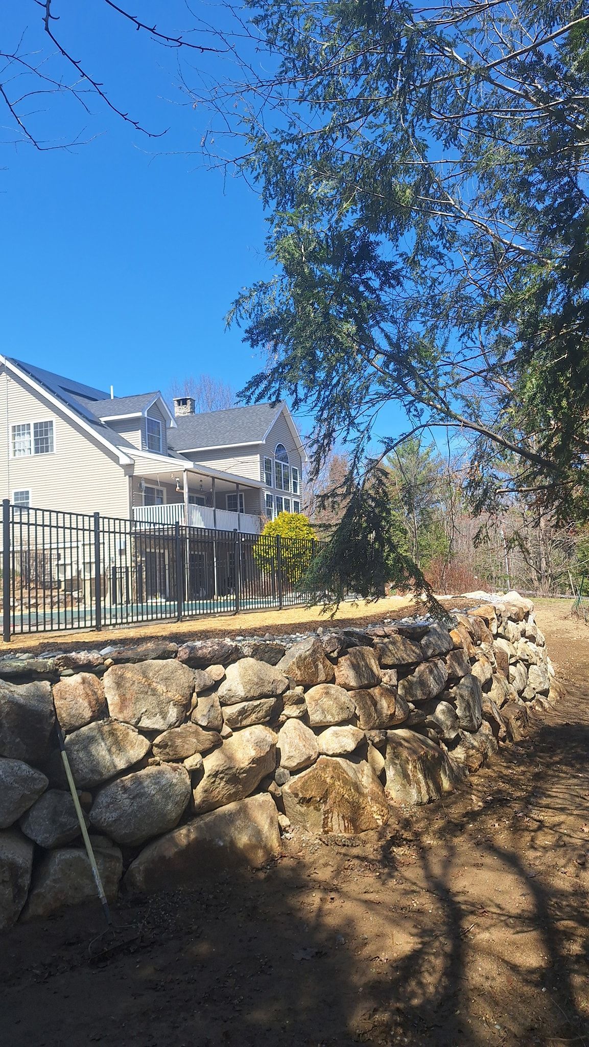 Stone retaining wall, fenced yard, trees, and houses under a blue sky.