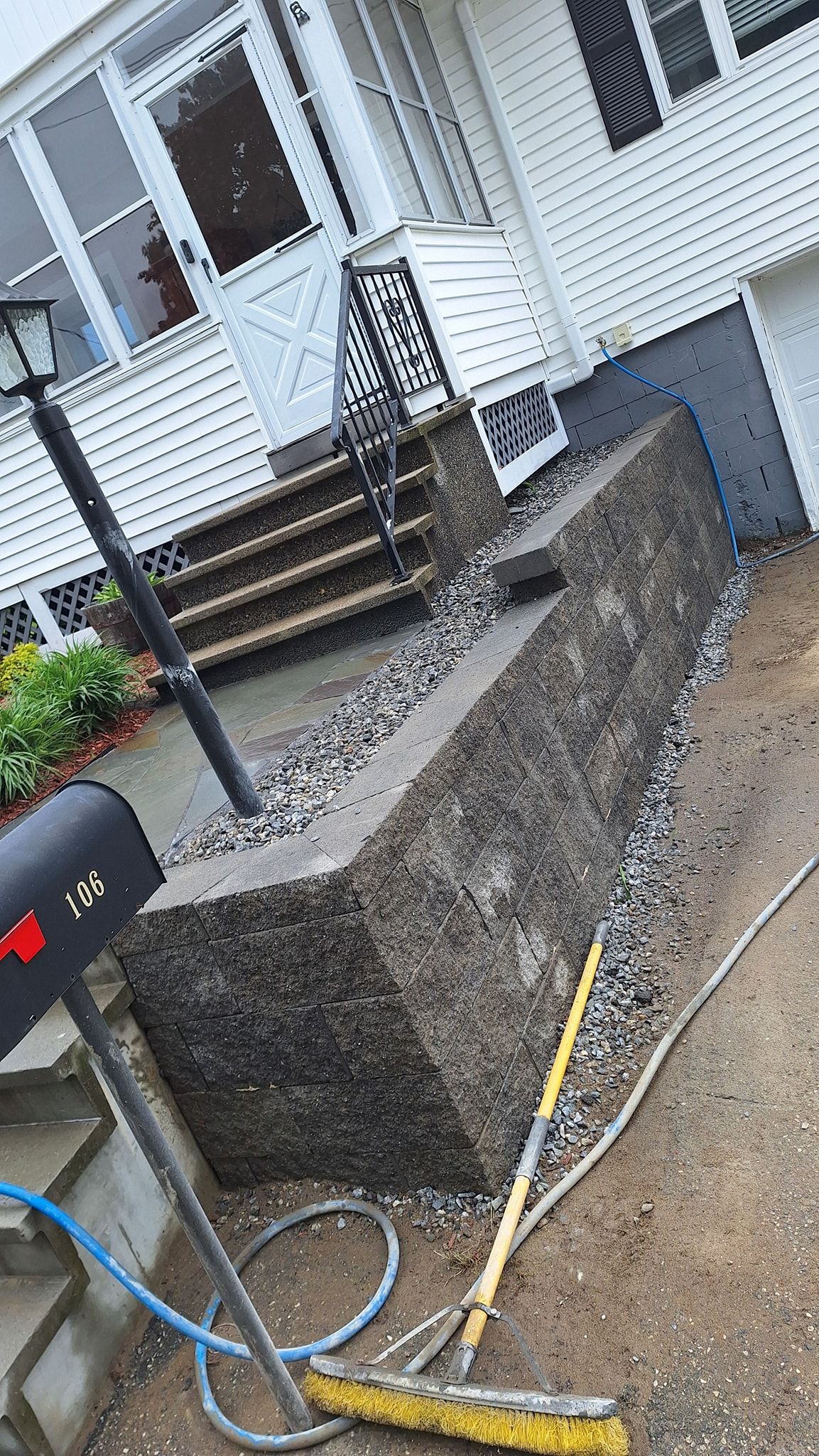 Concrete wall beside a house with a mailbox, steps, and gravel. A broom lies on the ground.