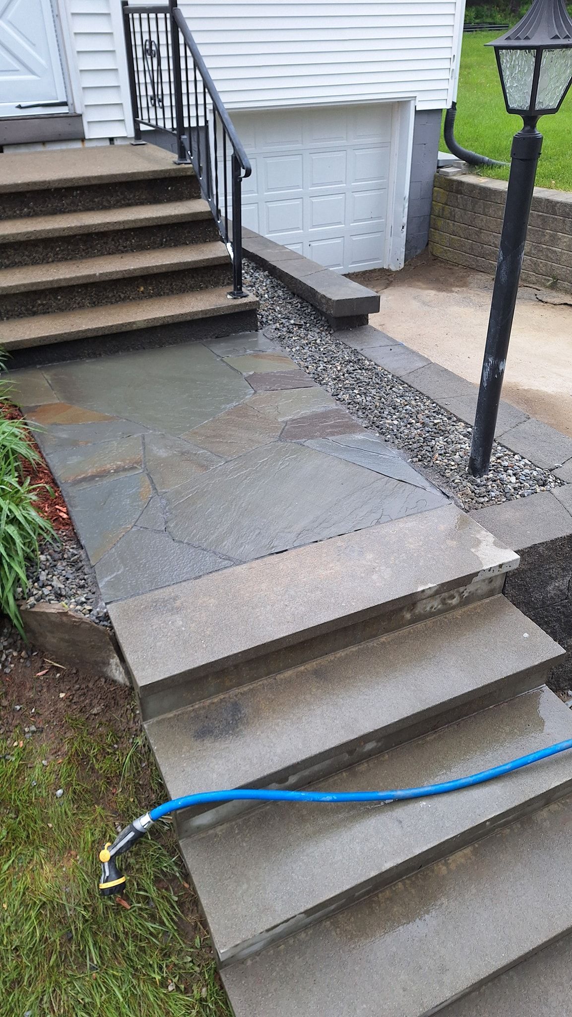 Concrete steps and walkway leading to a garage door; blue hose on steps, black railing, and landscaping.