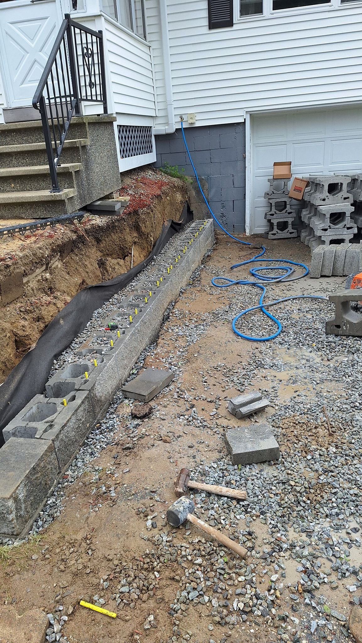 Construction site with concrete blocks, gravel, and dirt next to a house. Tools and blue tubing are visible.