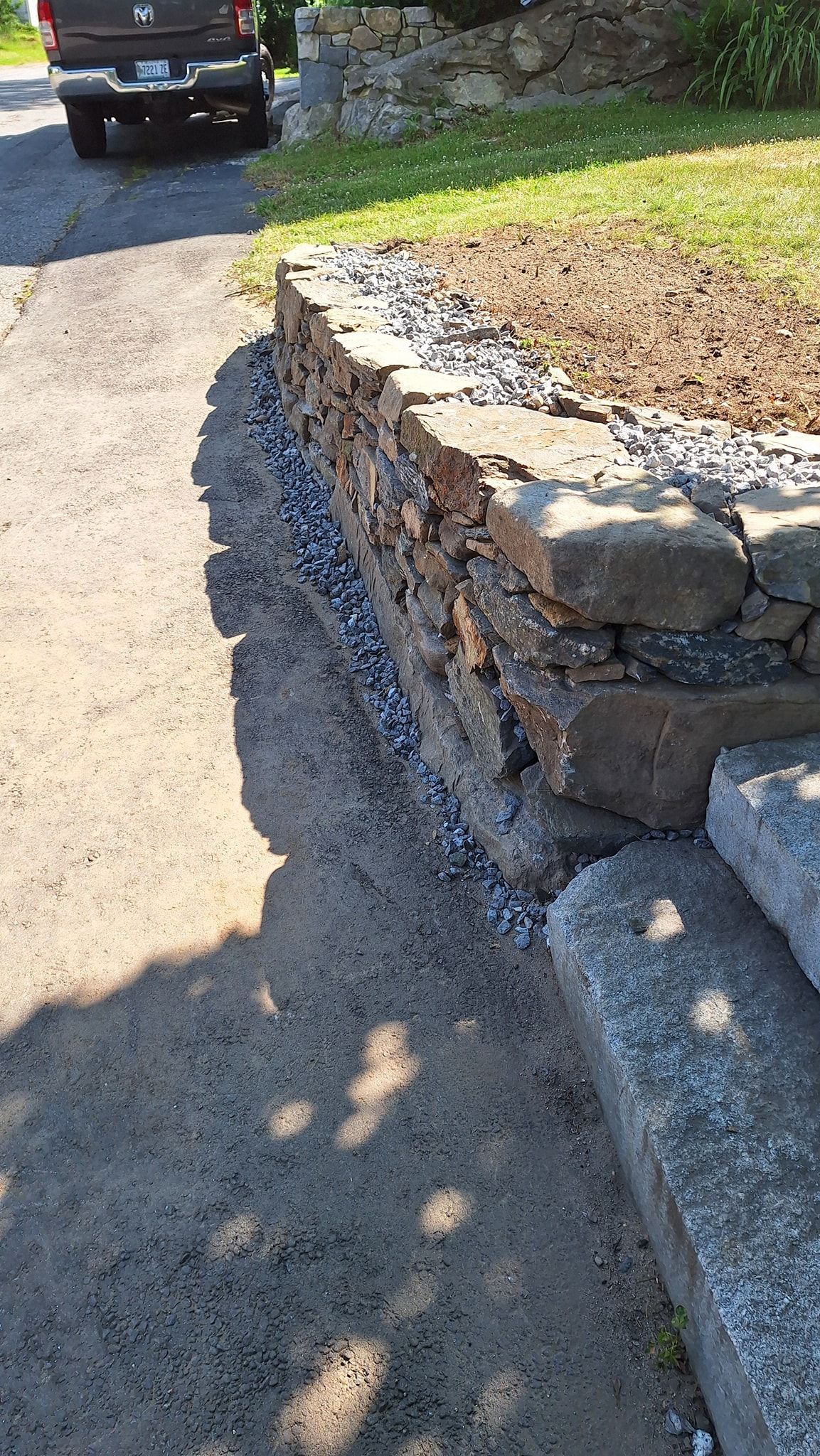Stone retaining wall with gravel base alongside a paved driveway.