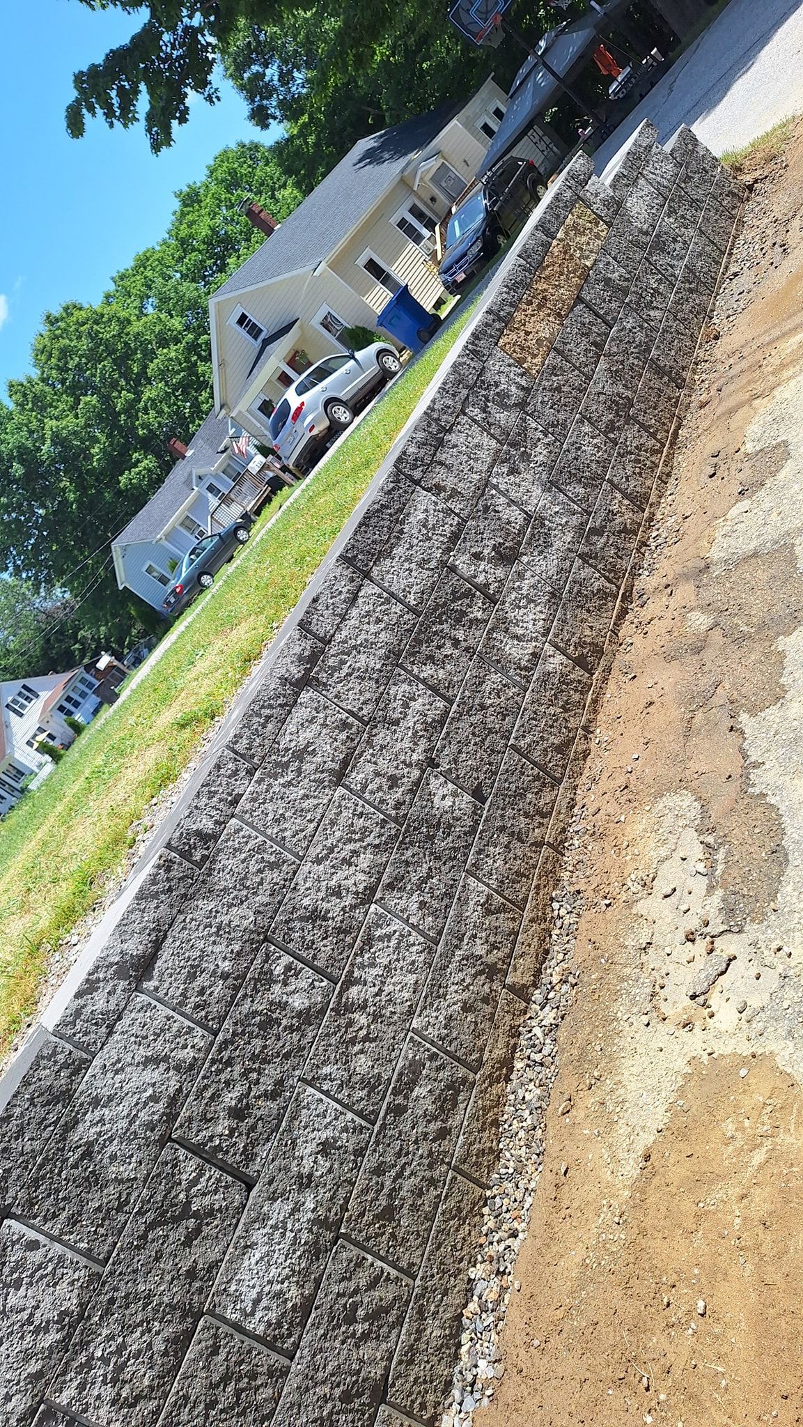 A gravel retaining wall alongside a road, with grass and houses visible in the background.