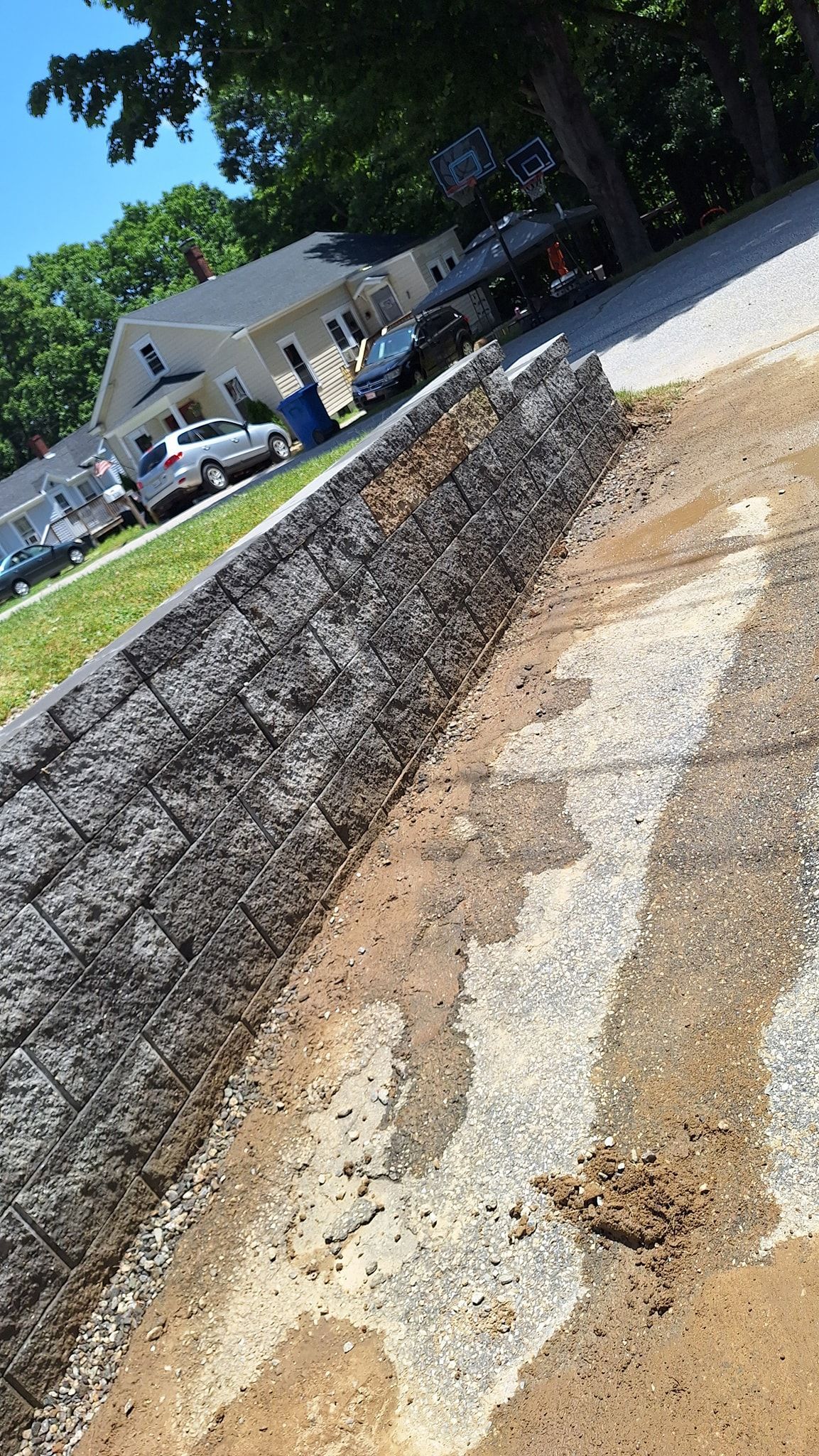 Gabion wall construction along a gravel driveway, with a house and parked cars in the background.