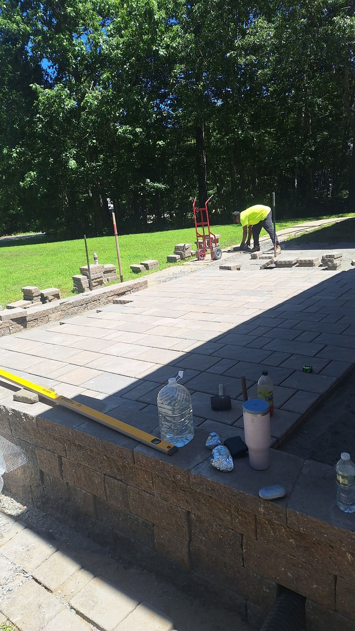Construction worker laying pavers on a patio outdoors; worker in a high-vis vest, tools, and supplies are present.