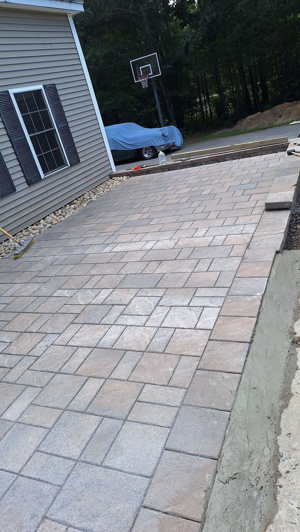 Paver walkway alongside a house with a gray and tan pattern; a car under a blue cover is in the background.