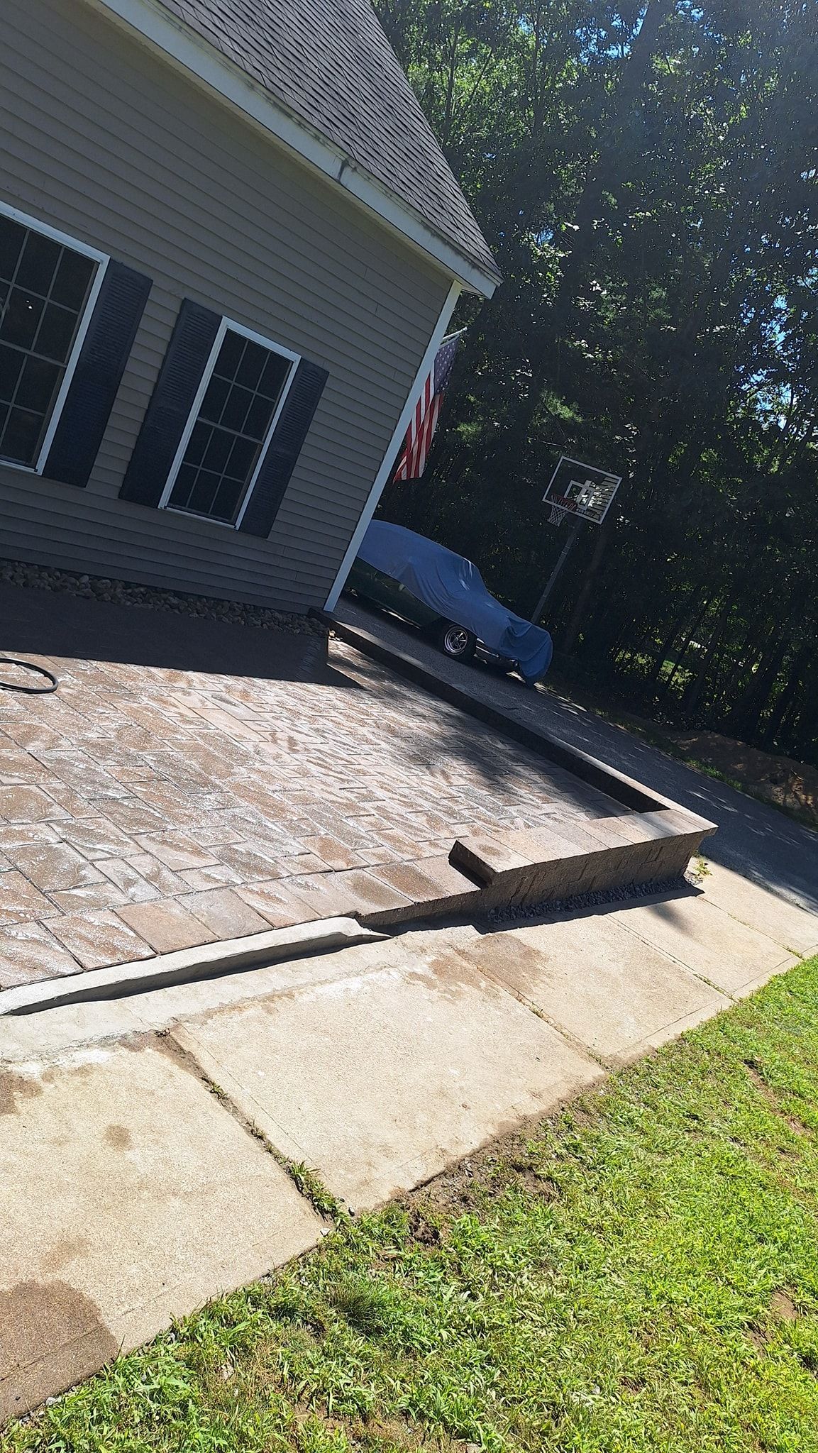 Patio with brick pavers and concrete border next to a house with dark shutters and green grass.