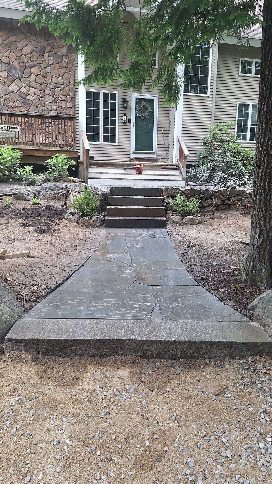 Stone path leads to a house with a green door and stacked firewood on the left.