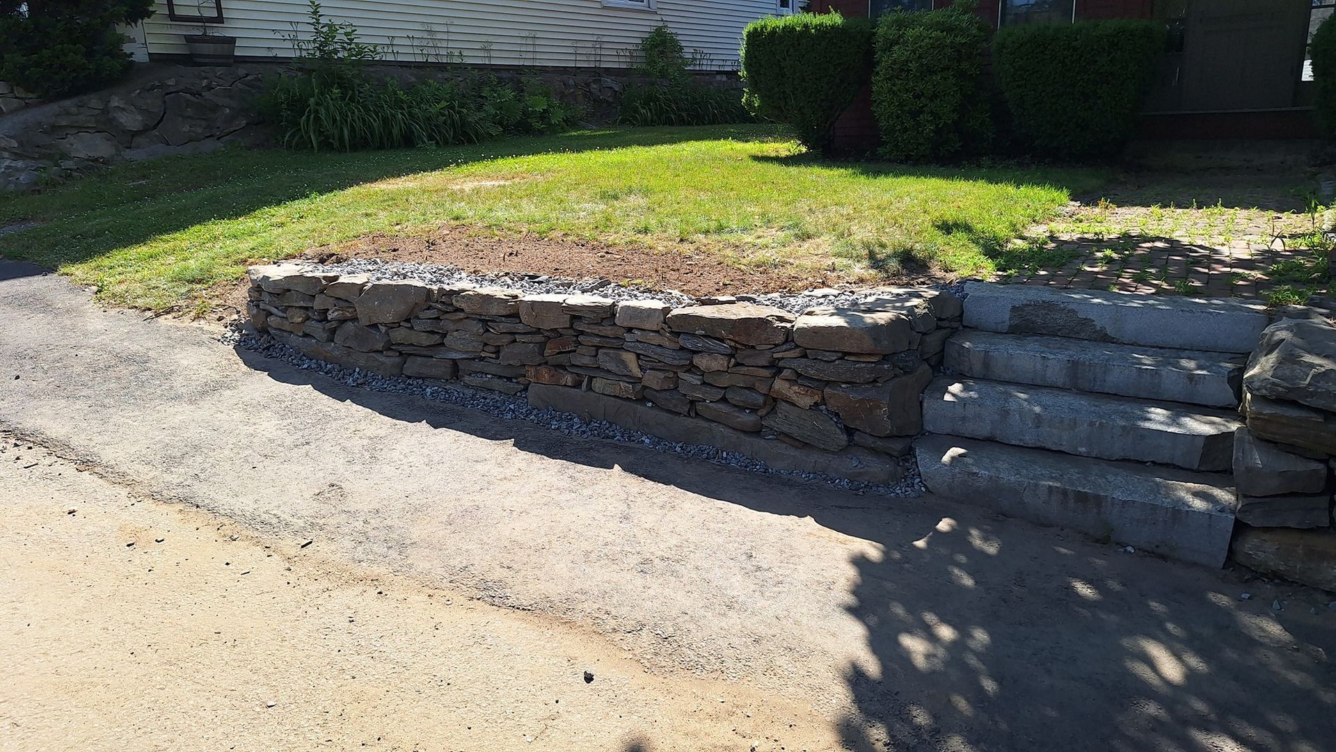 Stone retaining wall with steps next to a driveway and grass lawn.