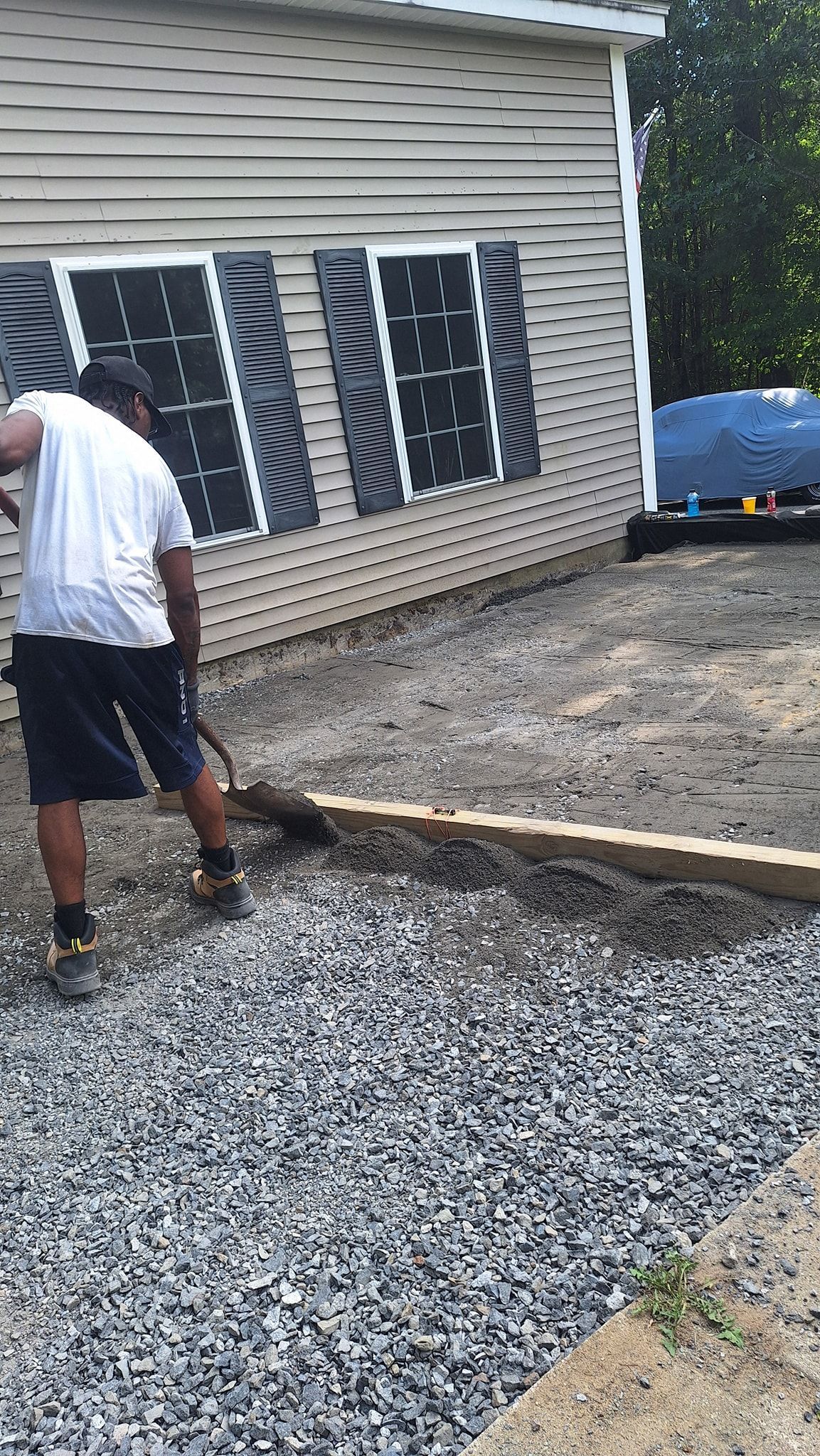 Man raking gravel near a building's side. Gravel path defined by wood. Building has closed shutters.
