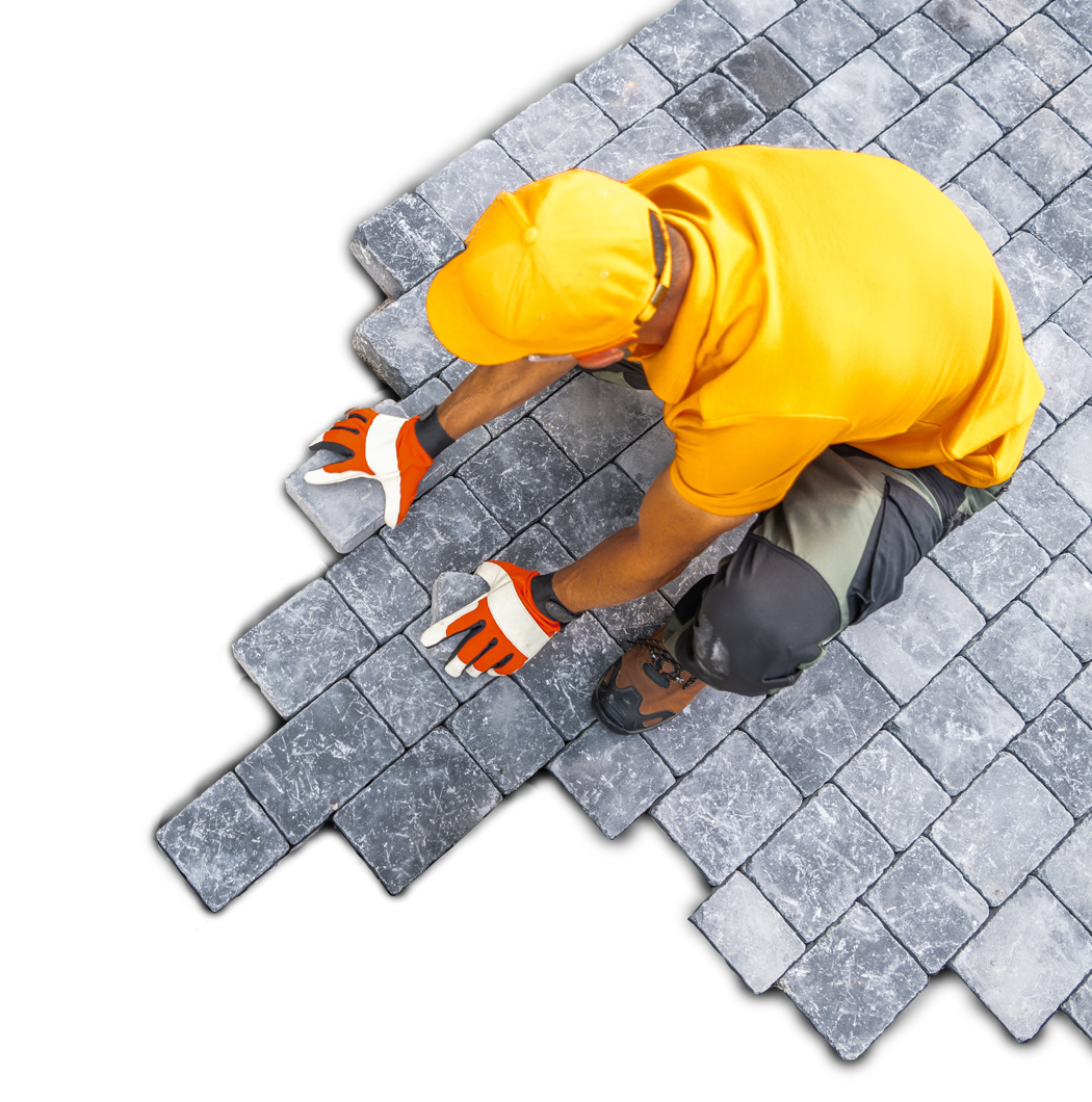 Worker laying gray paving stones, wearing yellow shirt, hat, and orange gloves.