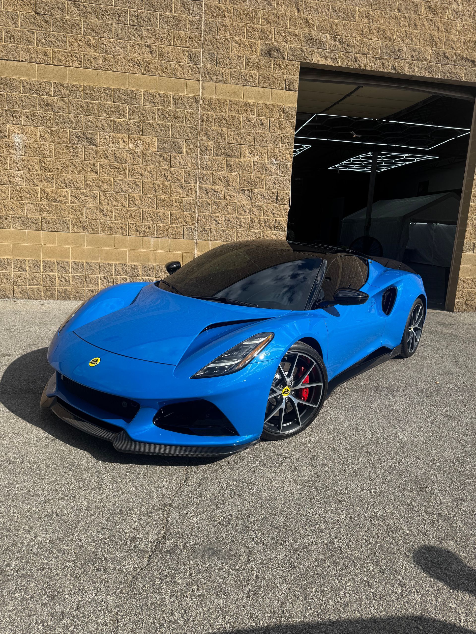 Blue Lotus Emira sports car parked in front of a brick building with an open garage.