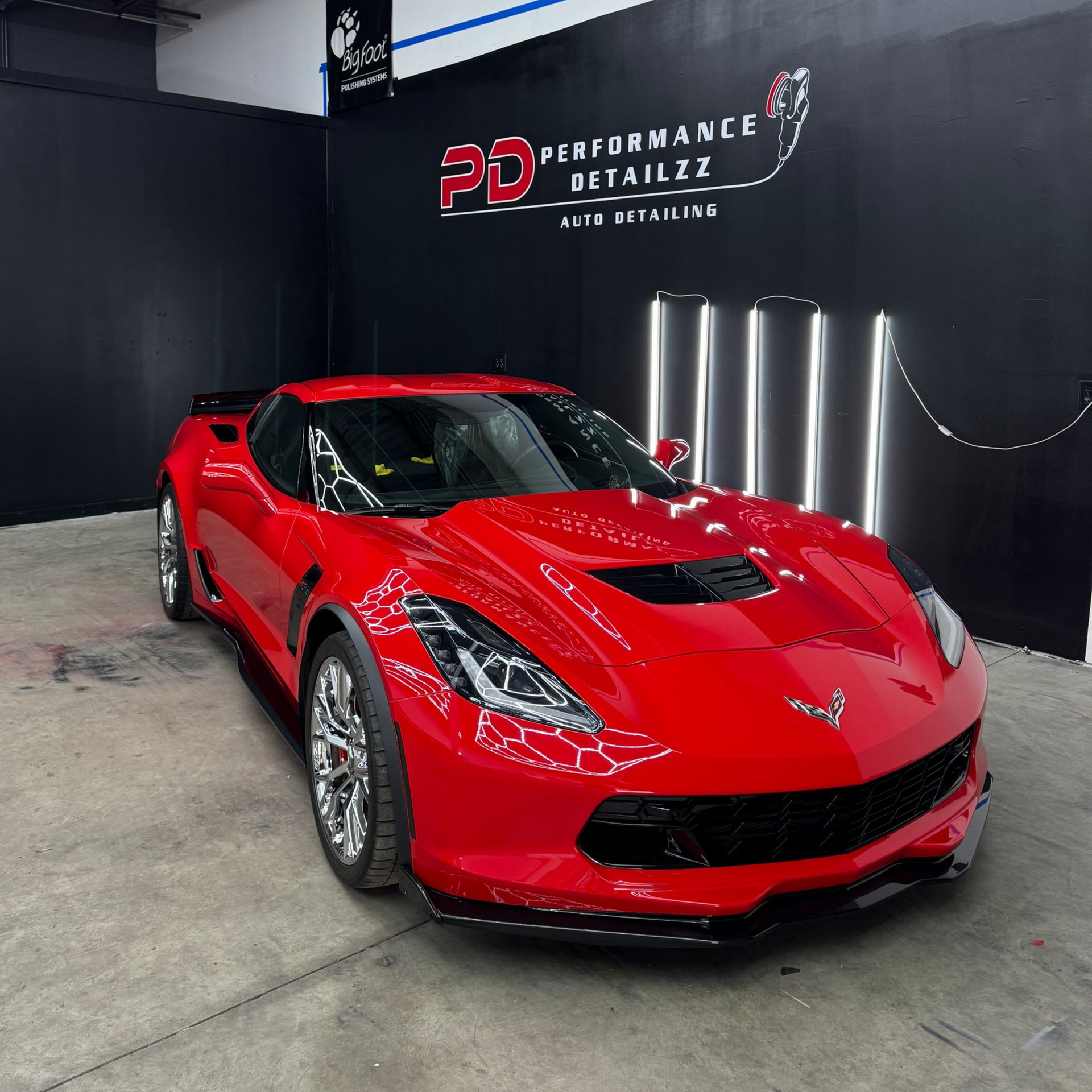 Red Corvette sports car parked in a detailing shop, black walls, and overhead lights.