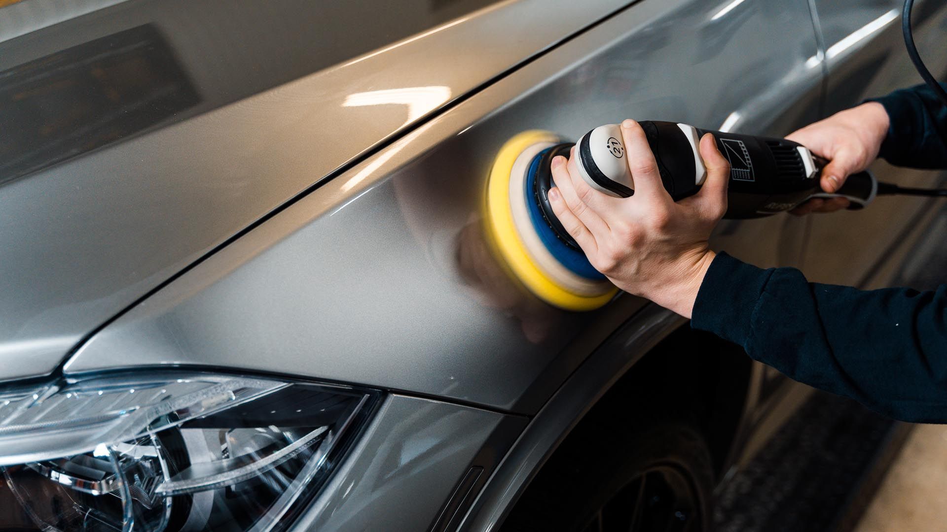 Person polishing gray car with an electric buffer in a garage.