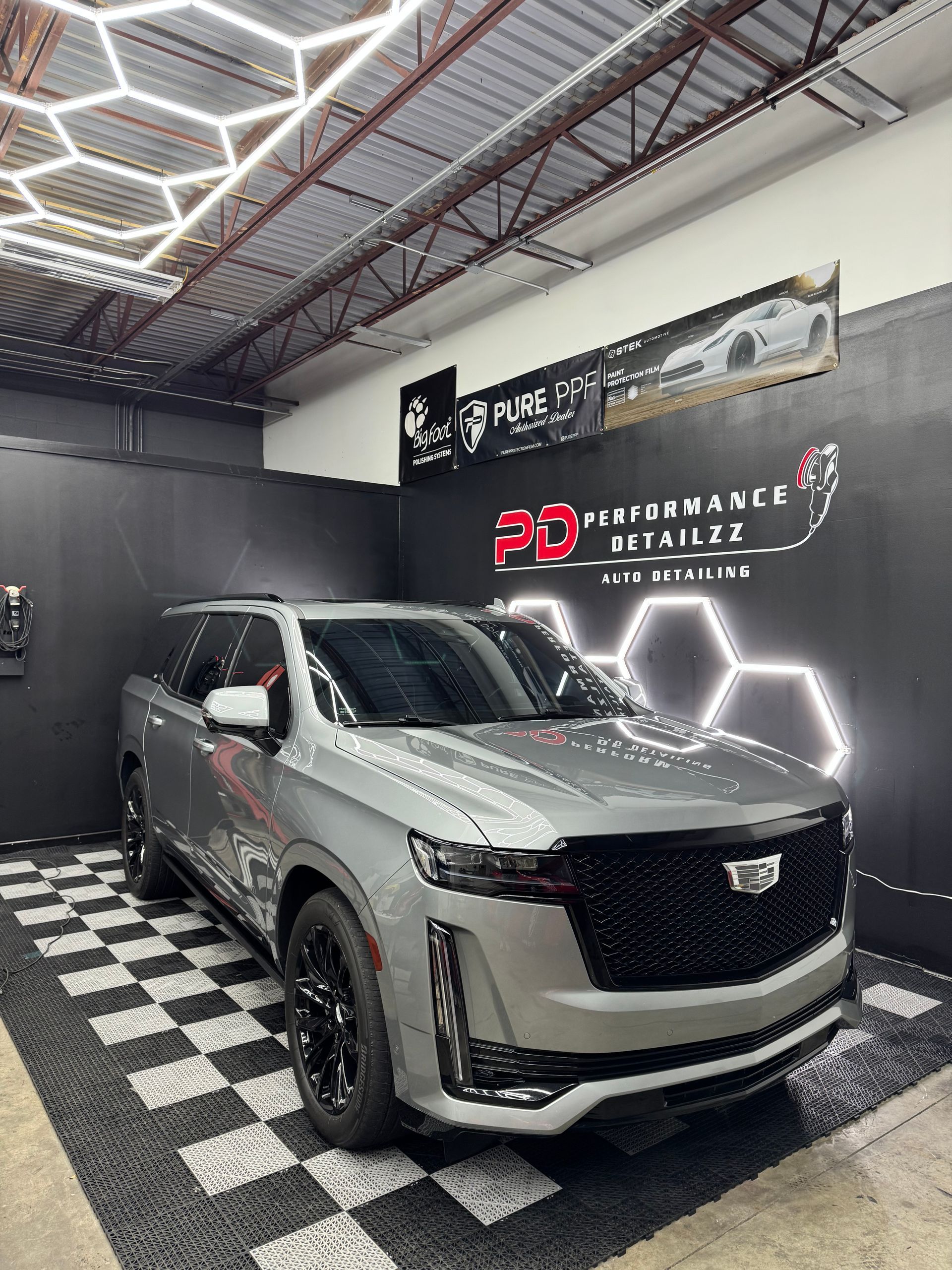 Silver Cadillac Escalade SUV in a detailing shop, black and white checkered floor, hexagonal lighting.