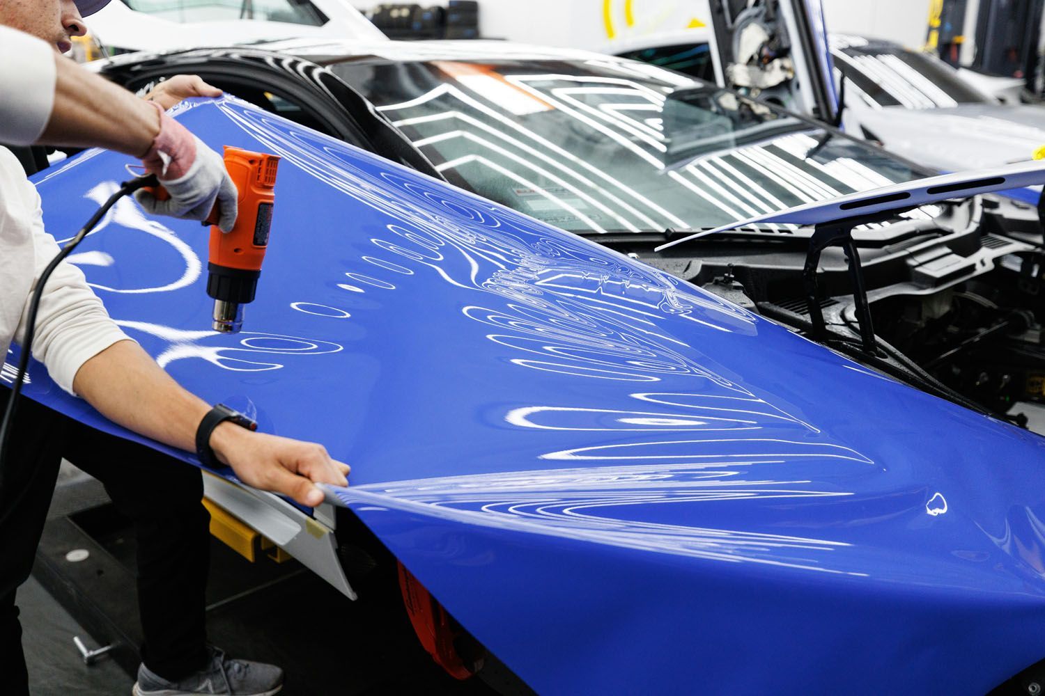 Person using a heat gun to apply blue vinyl wrap to a car hood in a workshop.