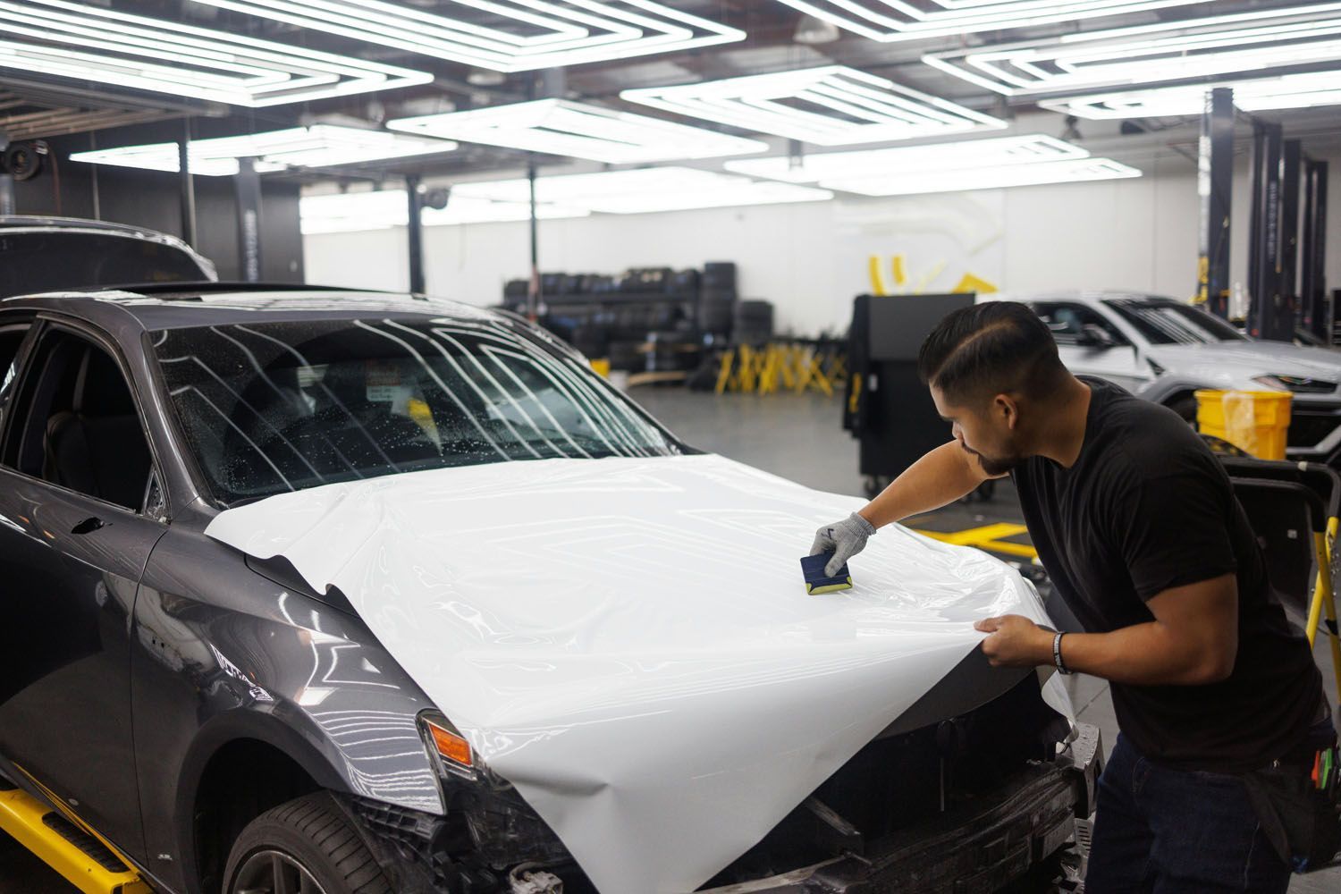 Man applying white vinyl wrap to the hood of a gray car in a garage.