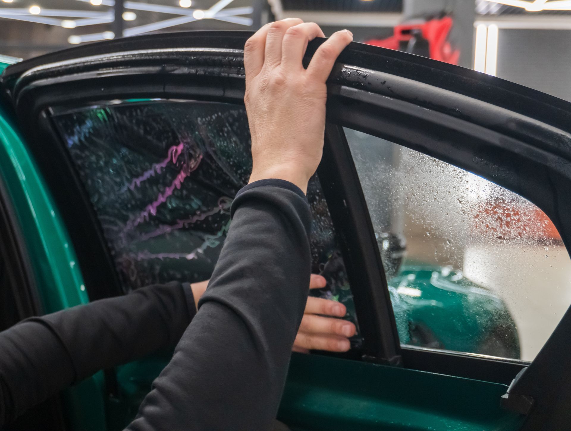 Hands installing tinted film on a car window in a garage.